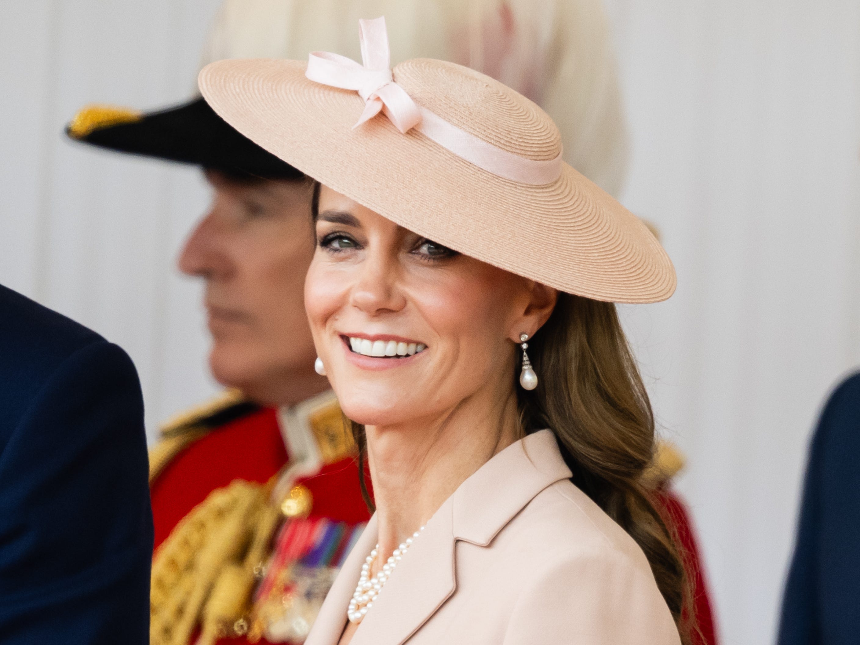 Kate Middleton smiles at a camera wearing a pink hat and jacket.