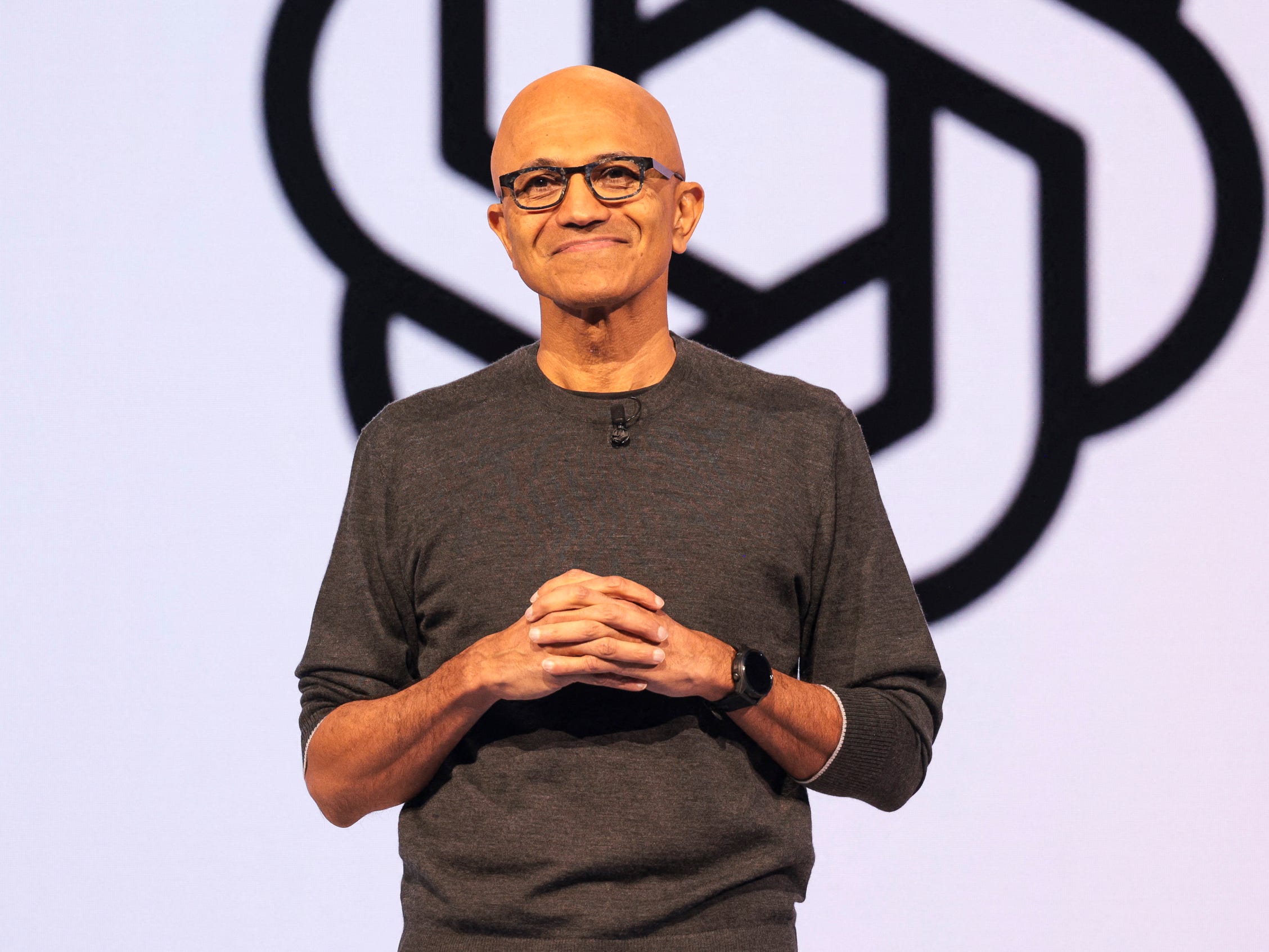 Satya Nadella wearing a brown sweater with his arms clasped in front of his chest, standing in front of a screen with the OpenAI logo.
