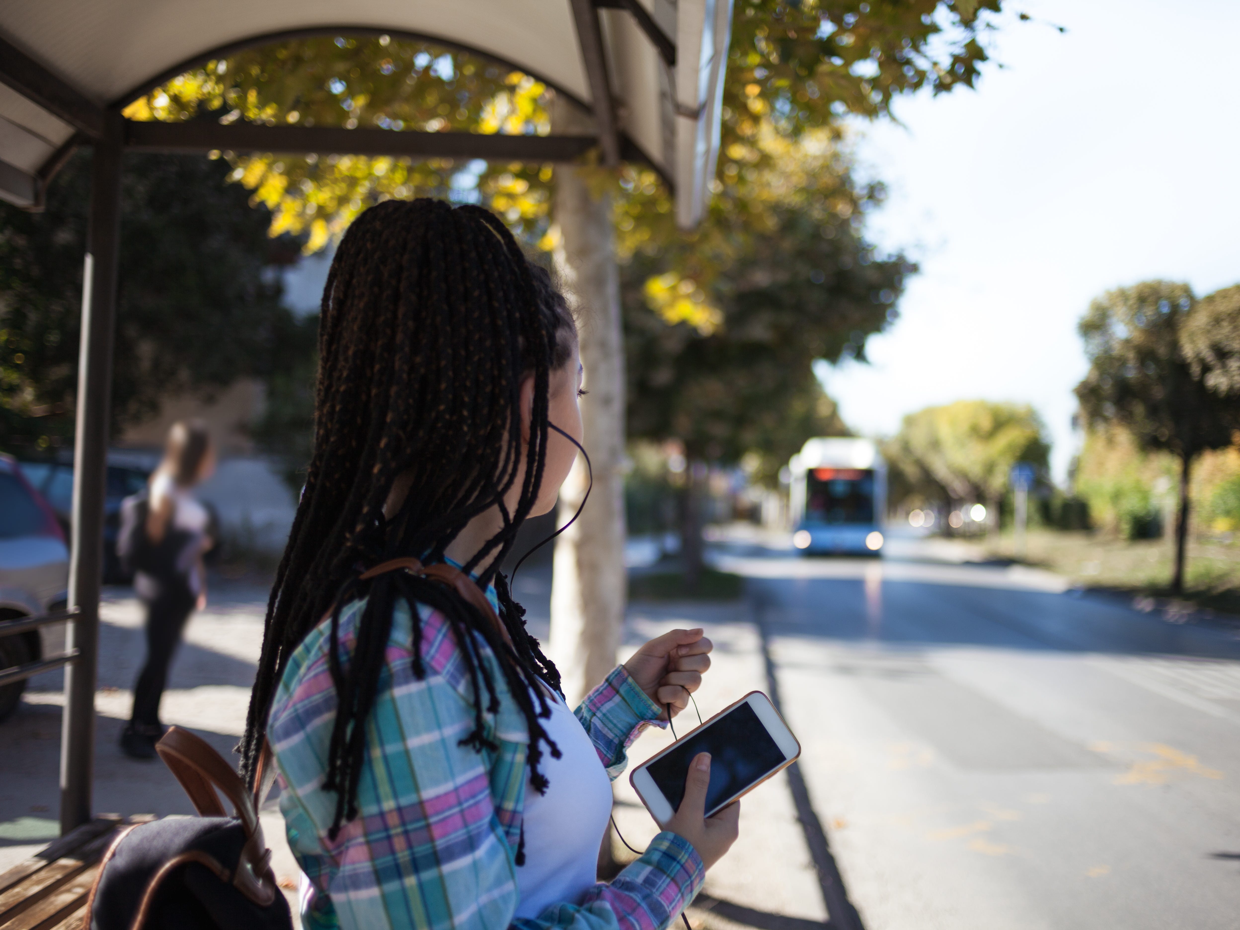 A teenage girl holding her phone and standing at a bus station waiting for the bus.