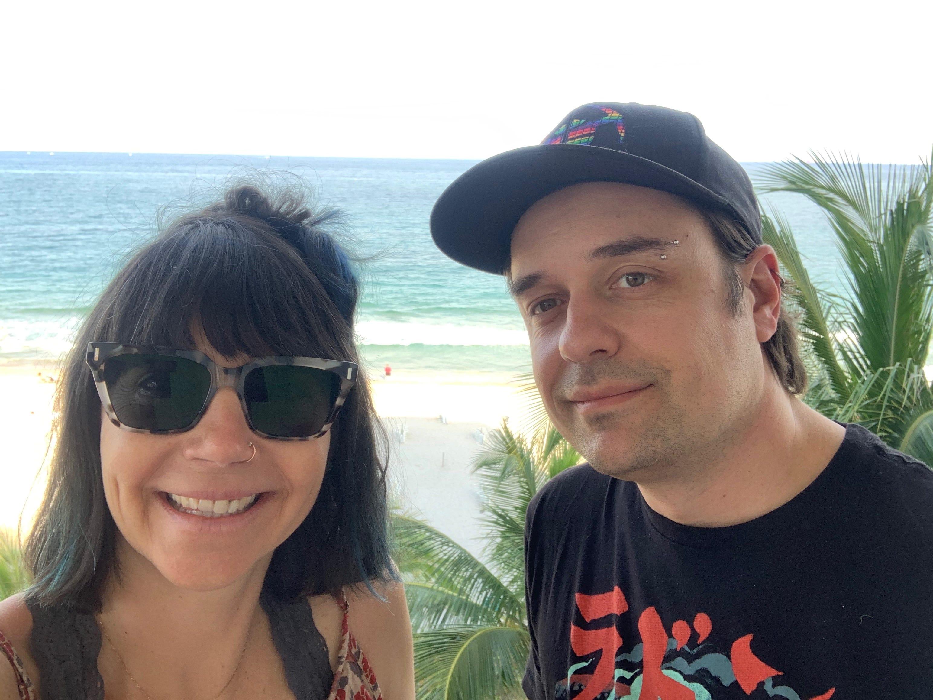 The author and her husband smiling and looking at the camera while standing on a beach. The ocean is behind them, she is wearing sunglasses and he has a hat on.