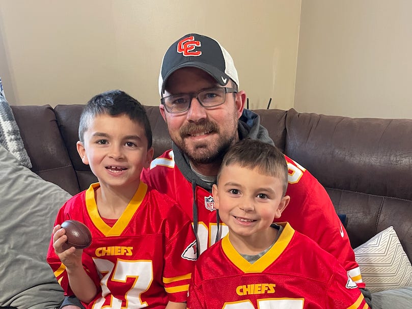 The author's husband with their two kids, all three wearing Chiefs jerseys.