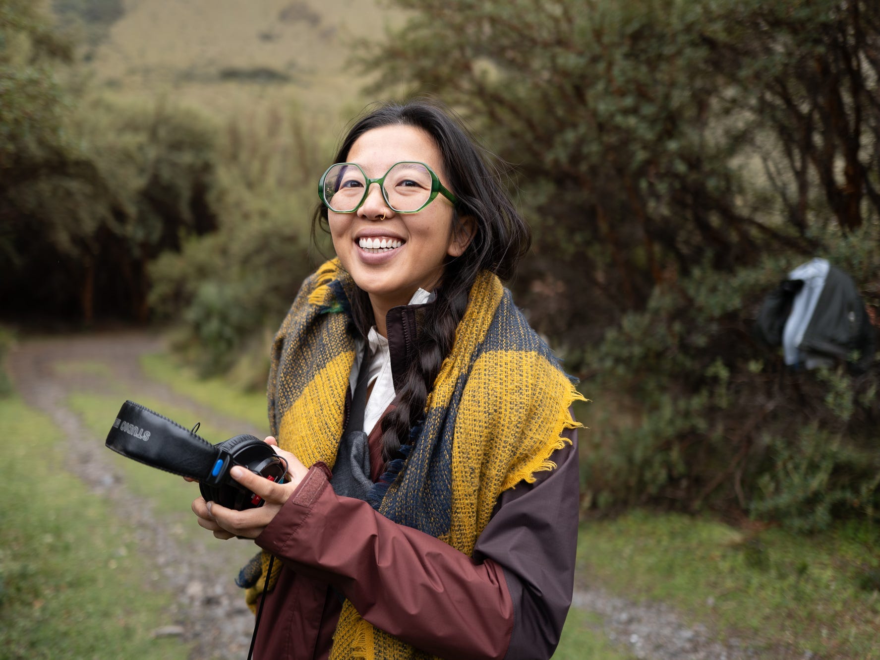 Ai Vuong, a woman wearing glasses, is standing holding earphones and standing outdoors with trees in the background.