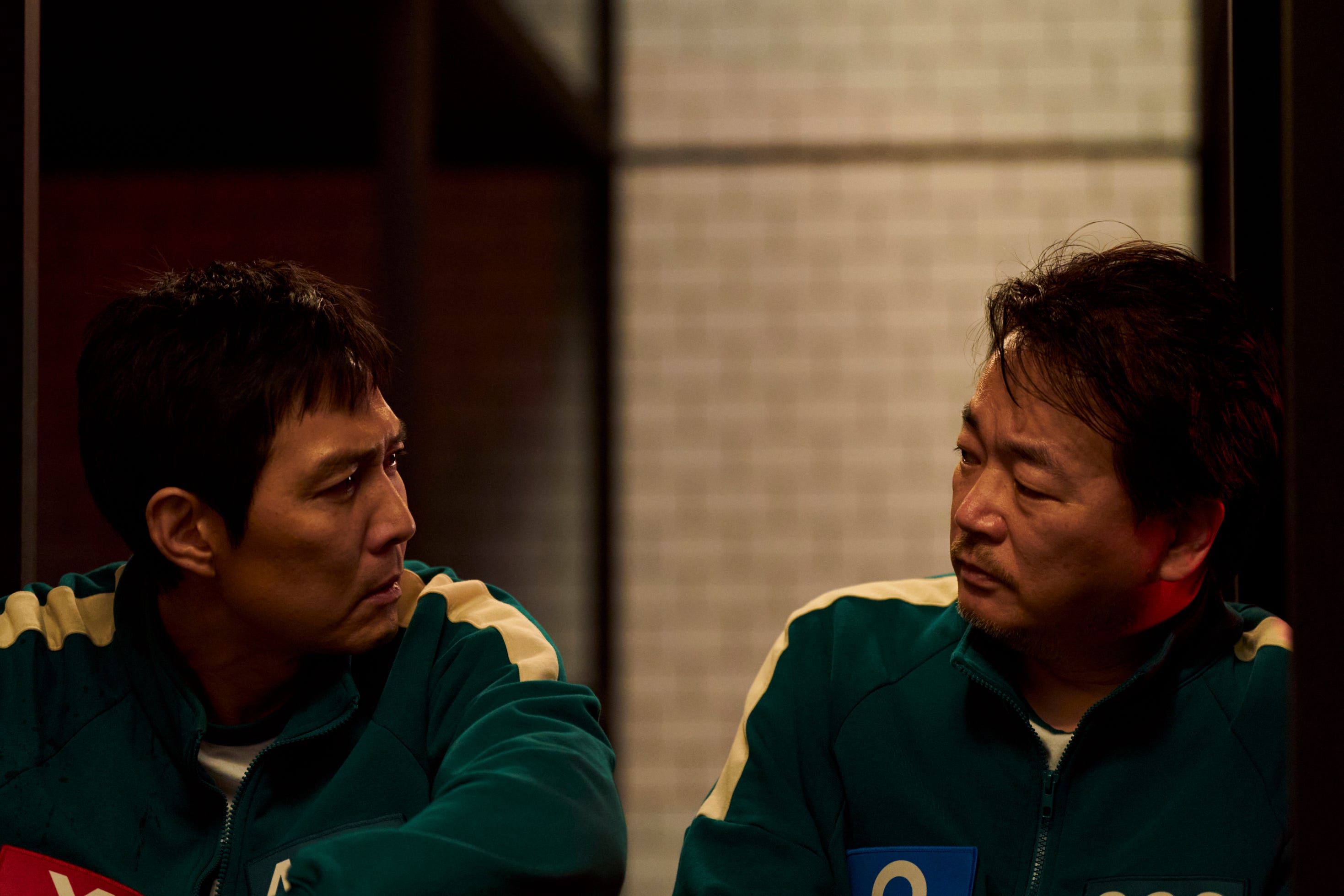 lee jung-jae and lee seo-hwan as gi-hun and jung-bae in season two of squid game. they're both middle aged men wearing green track suits, sitting together and looking at each other. gi-hun has a red x on his chest, while jung-bae has a blue circle