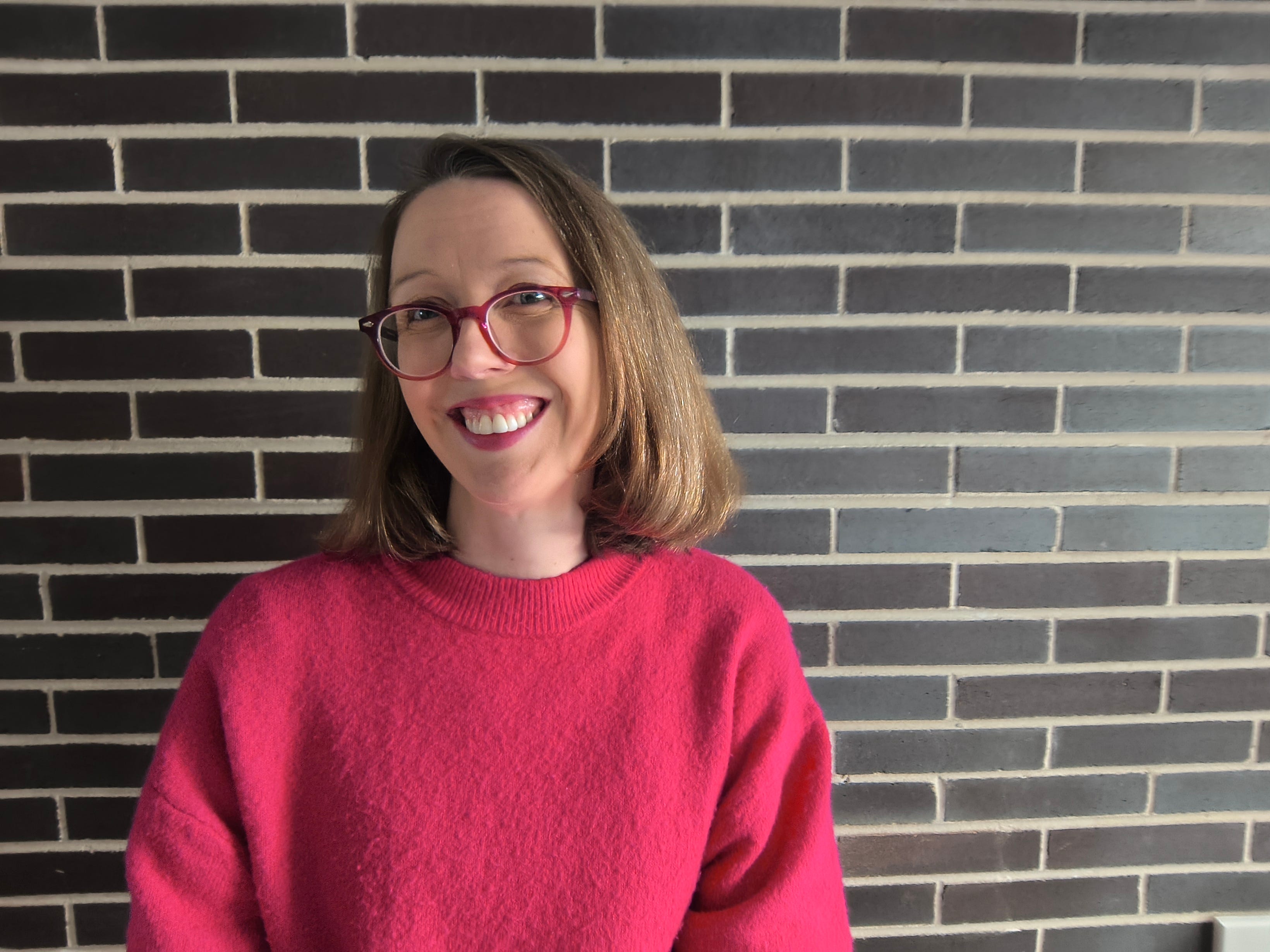 Dani Smith poses in front of a brick wall while wearing a reddish-pink sweater.