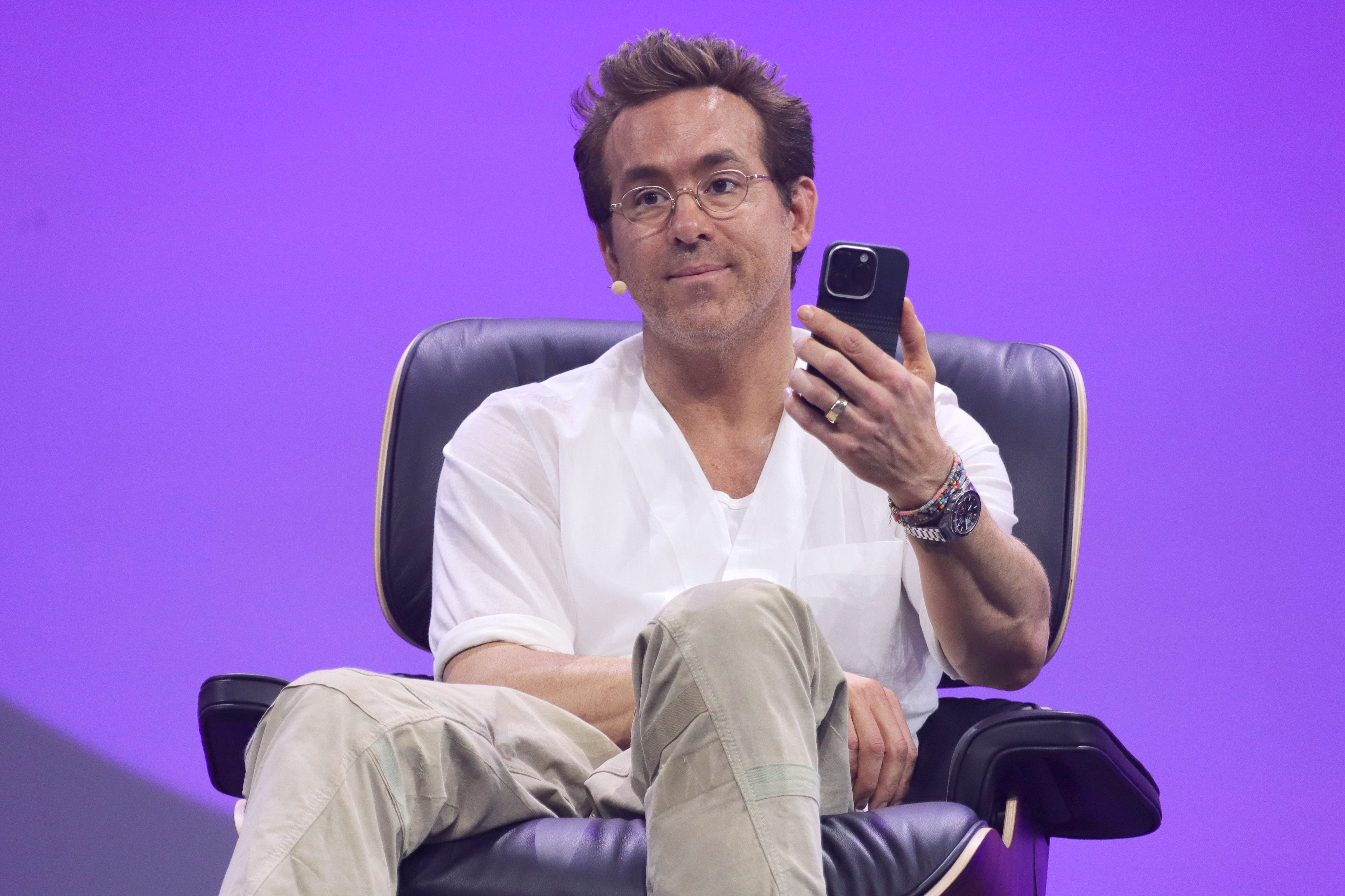 Ryan Reynolds sits on a stage holding a phone