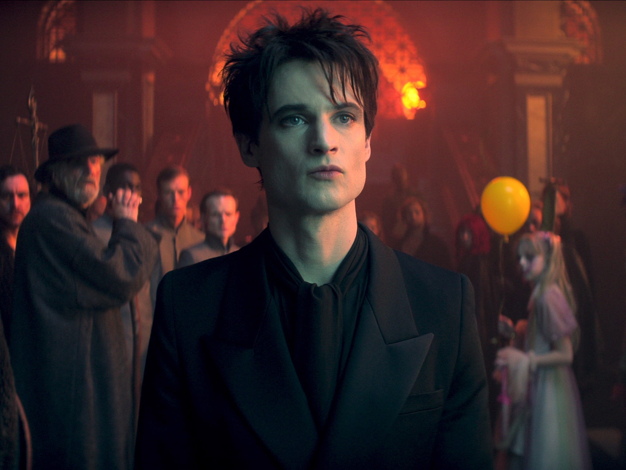 A pale man with messy black hair wears a black coat and necktie. He is surrounded by creepy clowns, an older bearded man with a beard, and slim men in high-collared jackets in a large ballroom with orange lighting in the background.