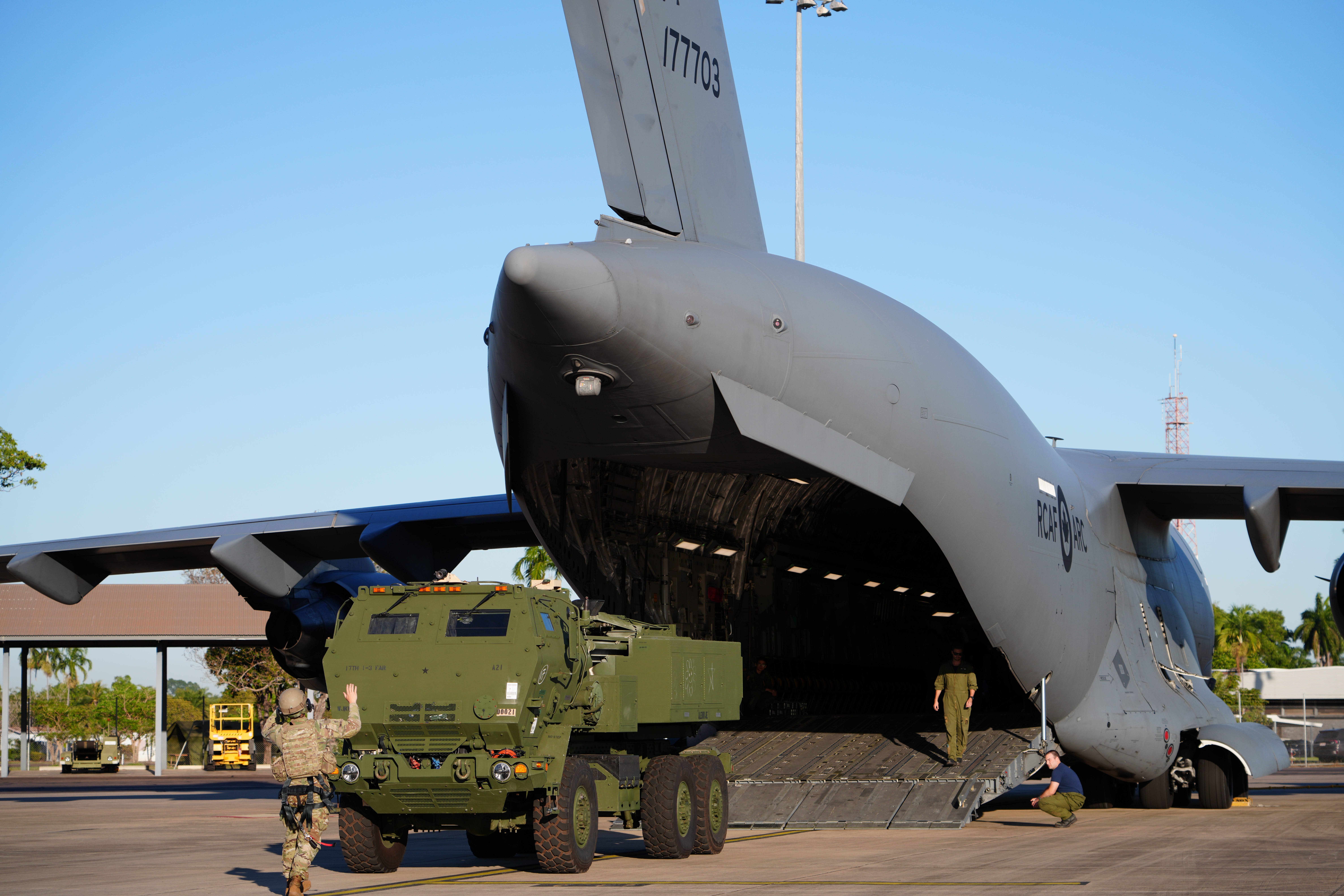A green truck drives off a large, grey military plane onto a tarmac. A man wearing a camouflage uniform stands in front of the truck, directing it. The sky is clear blue in the background.