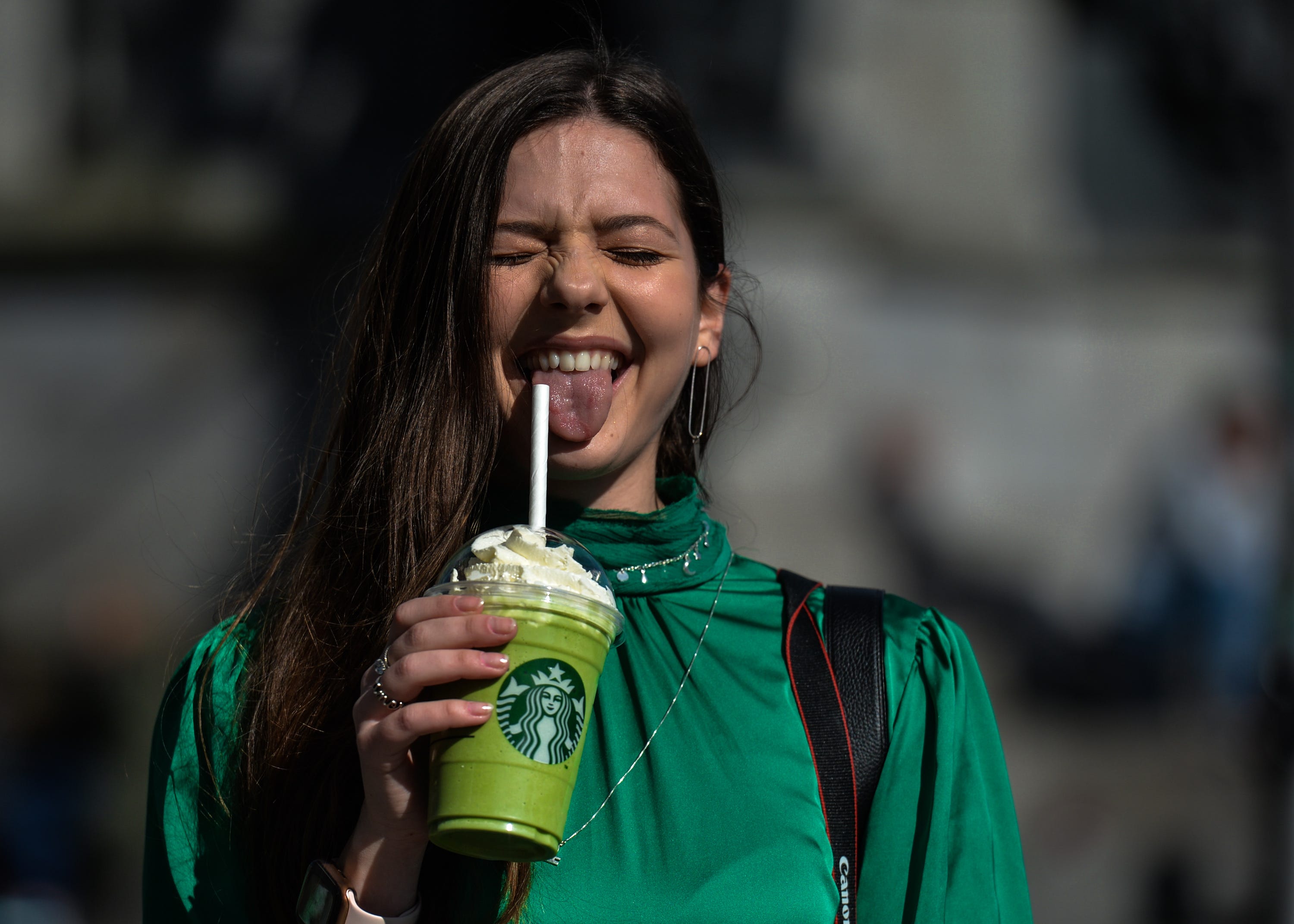 A woman smiles with her tongue out holding a green Starbucks drink in a clear plastic cup.