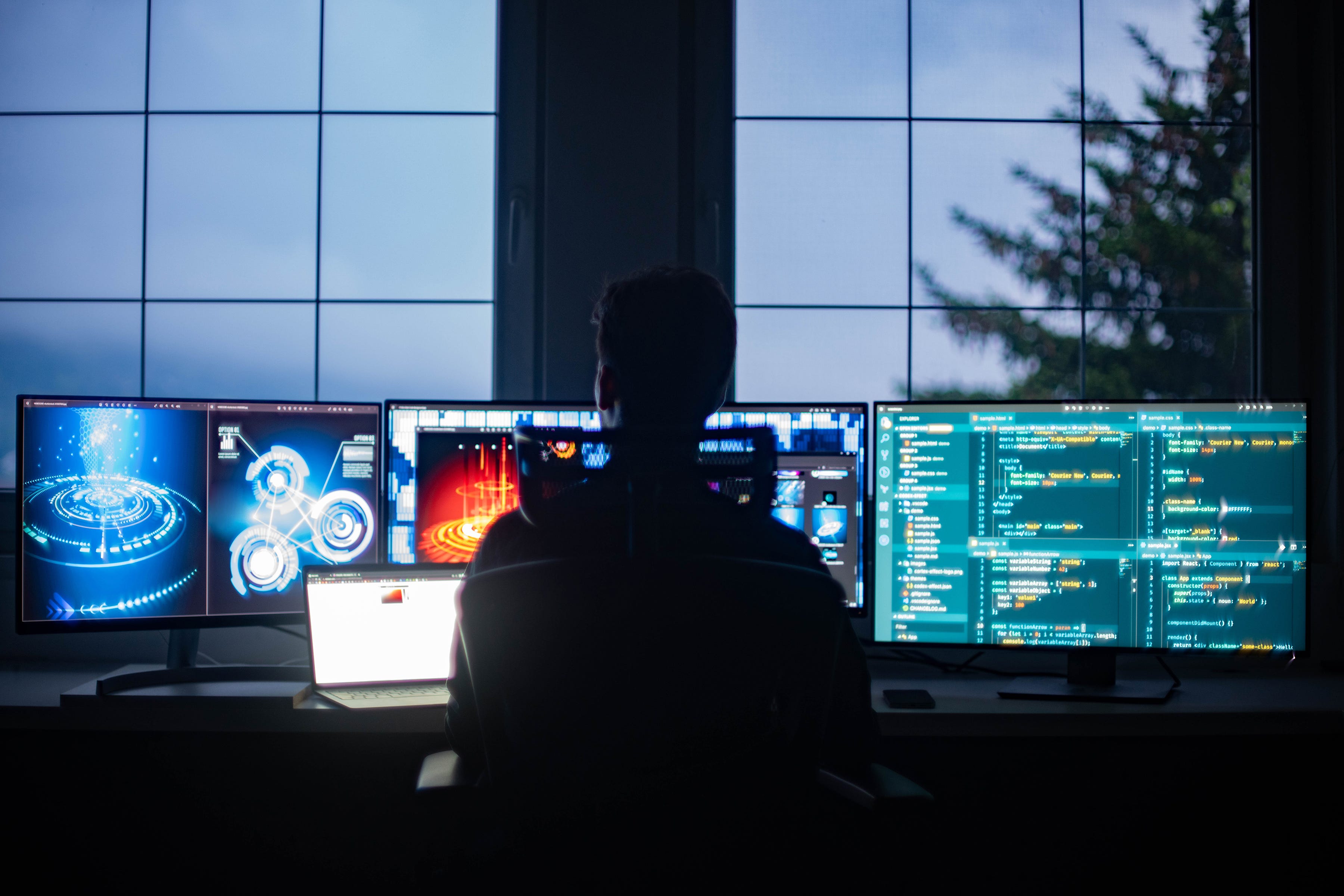 Man in a room at dusk at looking at multiple computer screens.