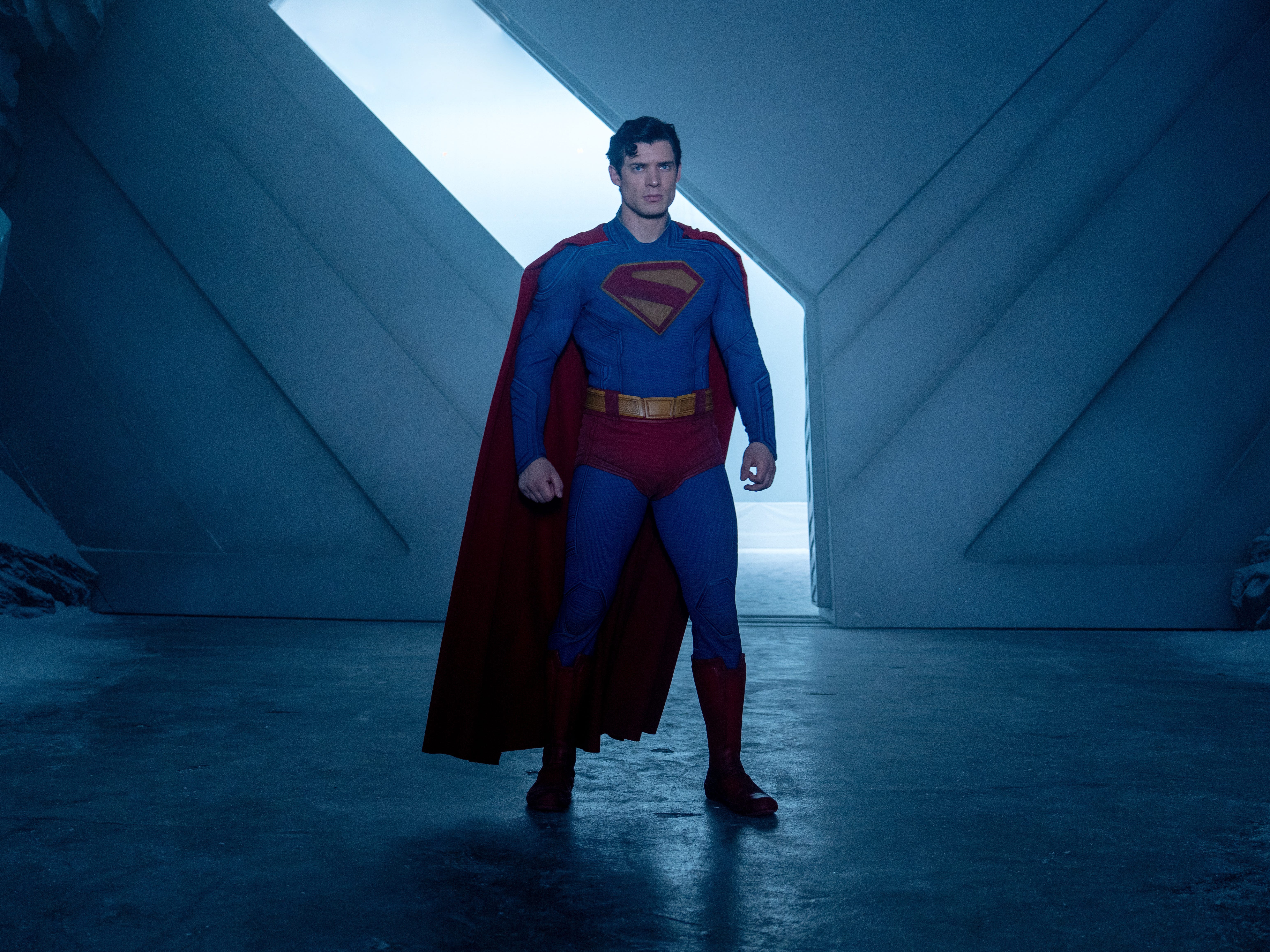 A man with black hair stands in an icy room with a large metal door behind him. He's wearing a blue costume with red trunks, a red cape, red boots and a red S on his chest.