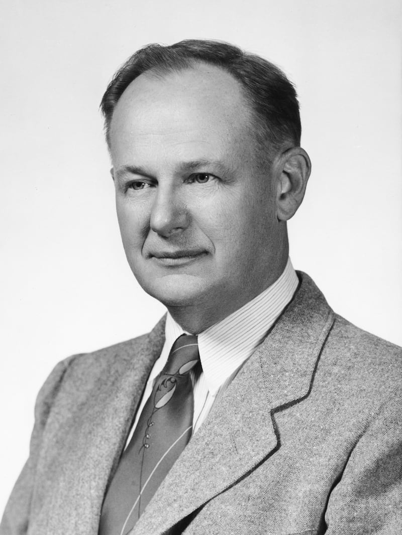 A portrait of a balding man, wearing a wide-lapeled light-colored jacket and tie. The portrait is black and white