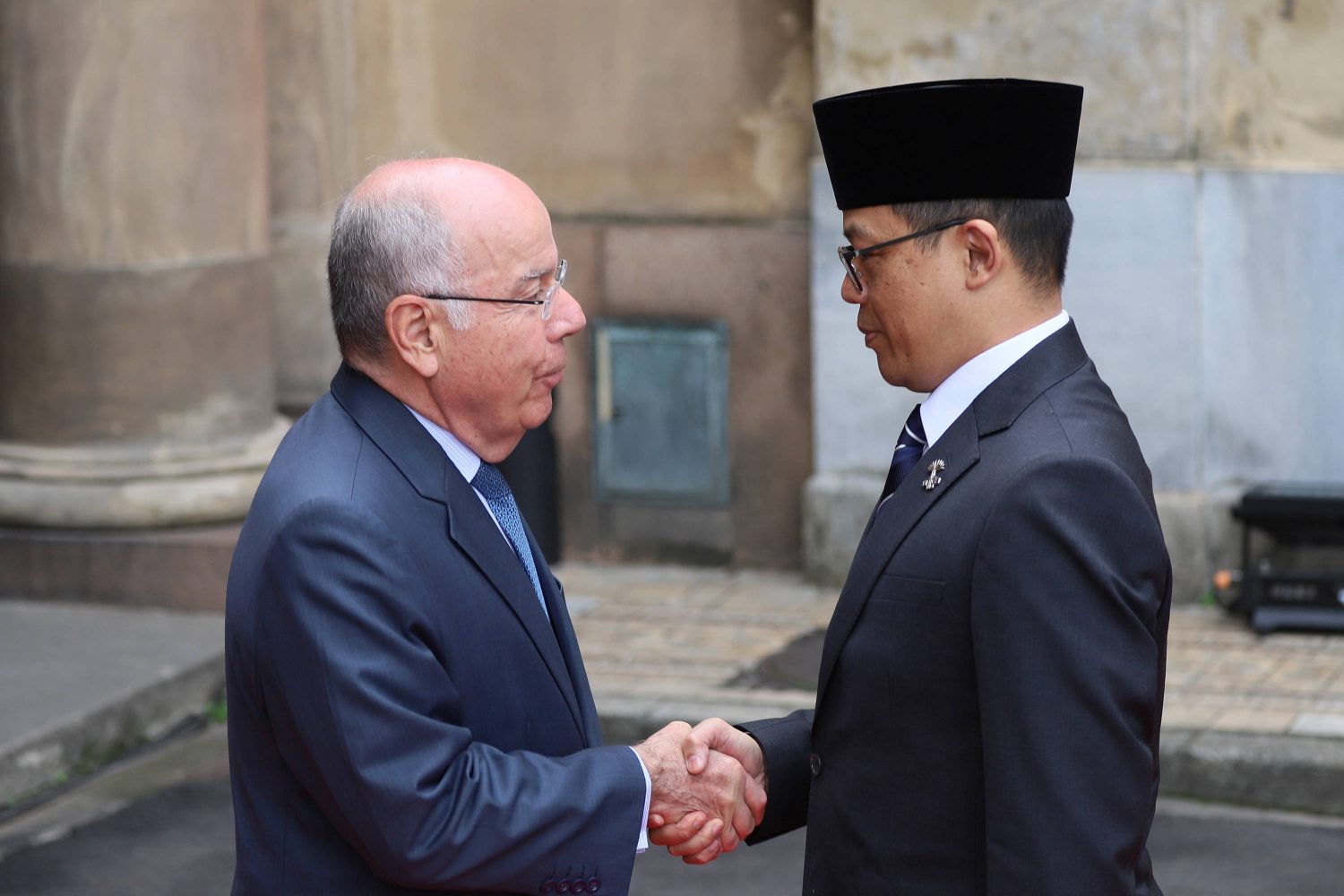 Two men in suits, shown from the side, shake hands.