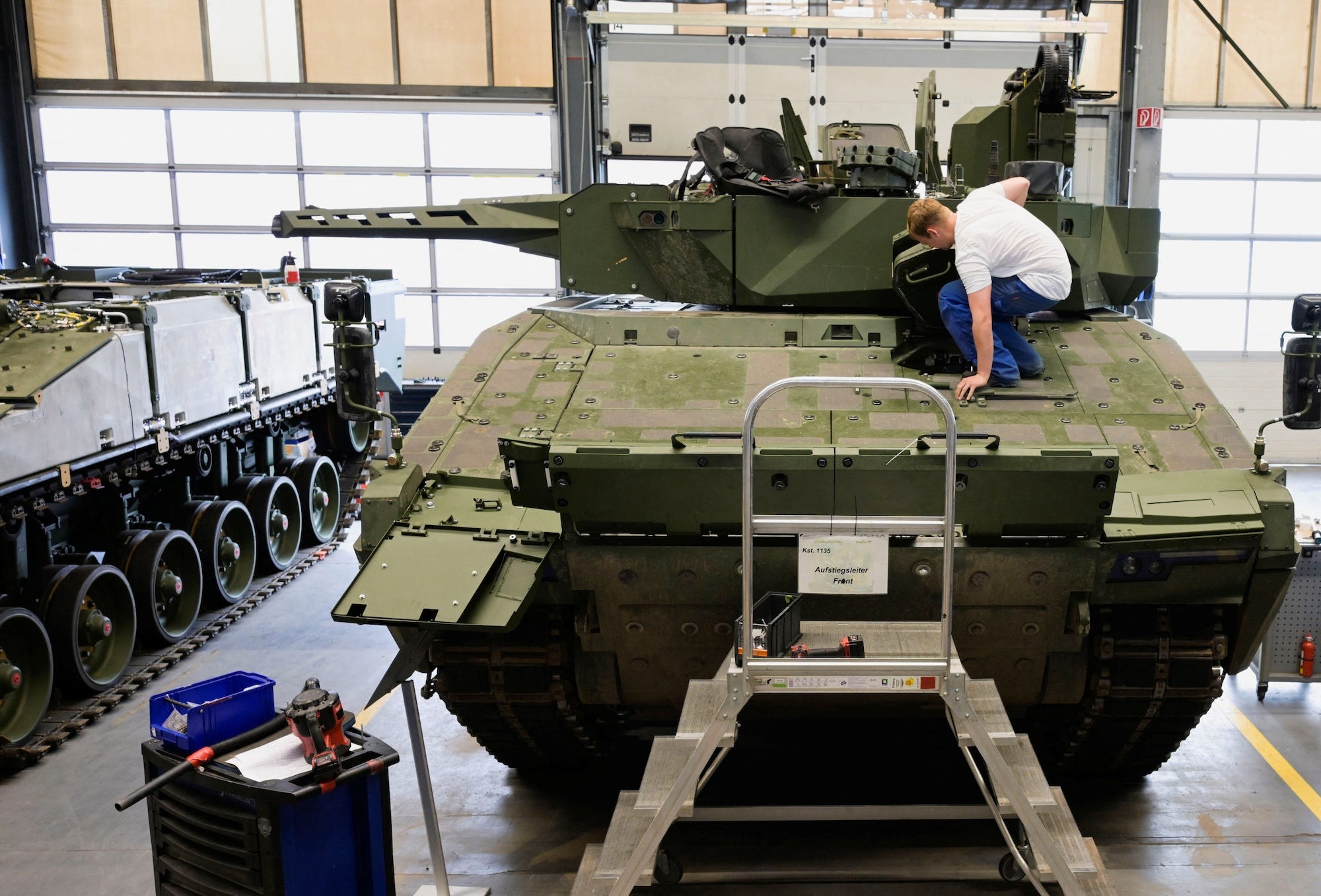 A man in a blue trousers and white t-shirt sqauts in front of a partially built green armored vehicle inside a factory room