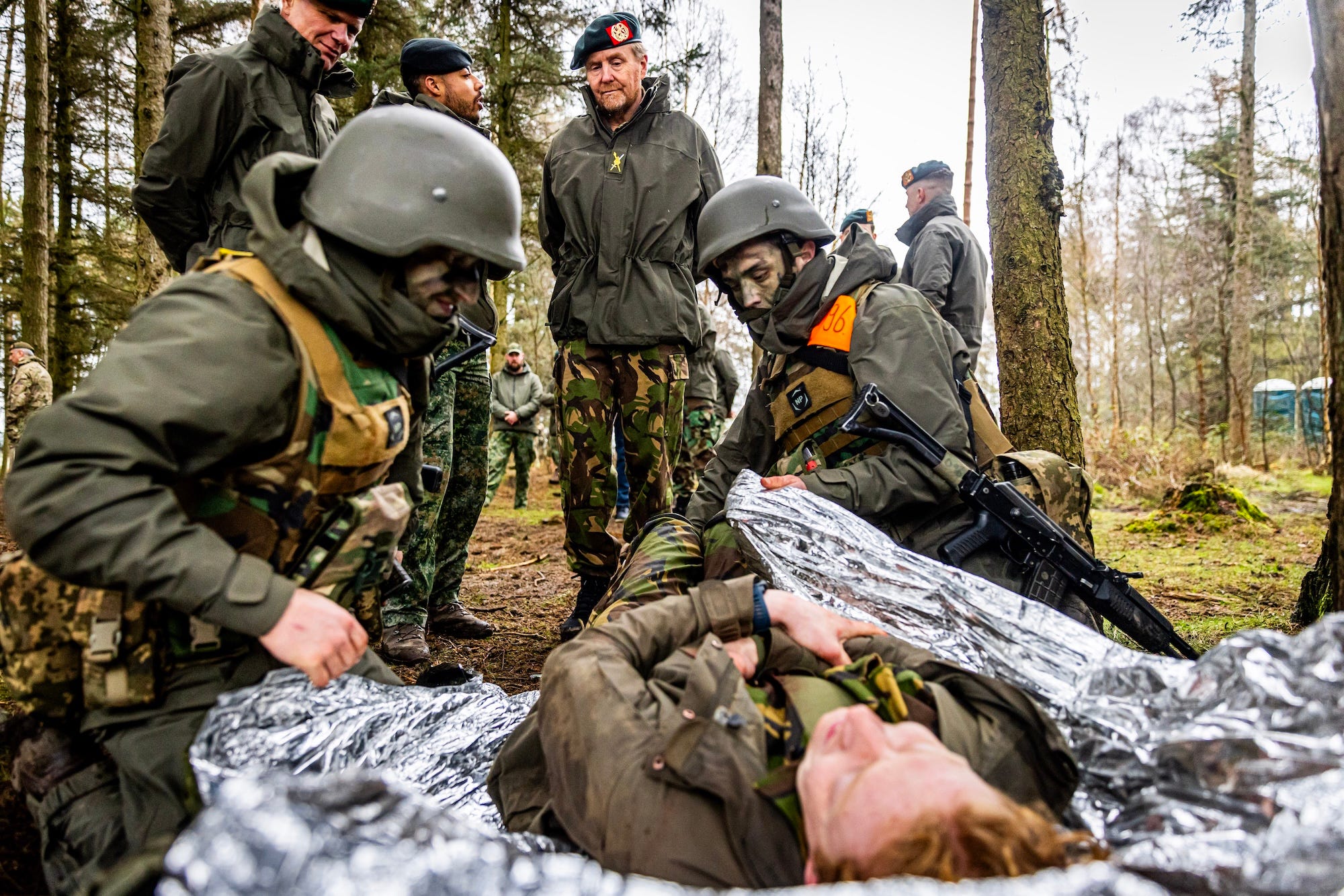 Two men in camouflage gear and helmets kneel on either side of a man lying on the ground and wrap him in a foil blanket as other men in military gear watch on, all surrounded by trees