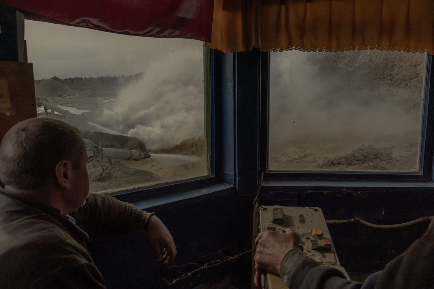 Two people are seen from behind as they sit at behind a dusty window, operating switchboard controls while watching a large piece of mining equipment kick up dust outside.