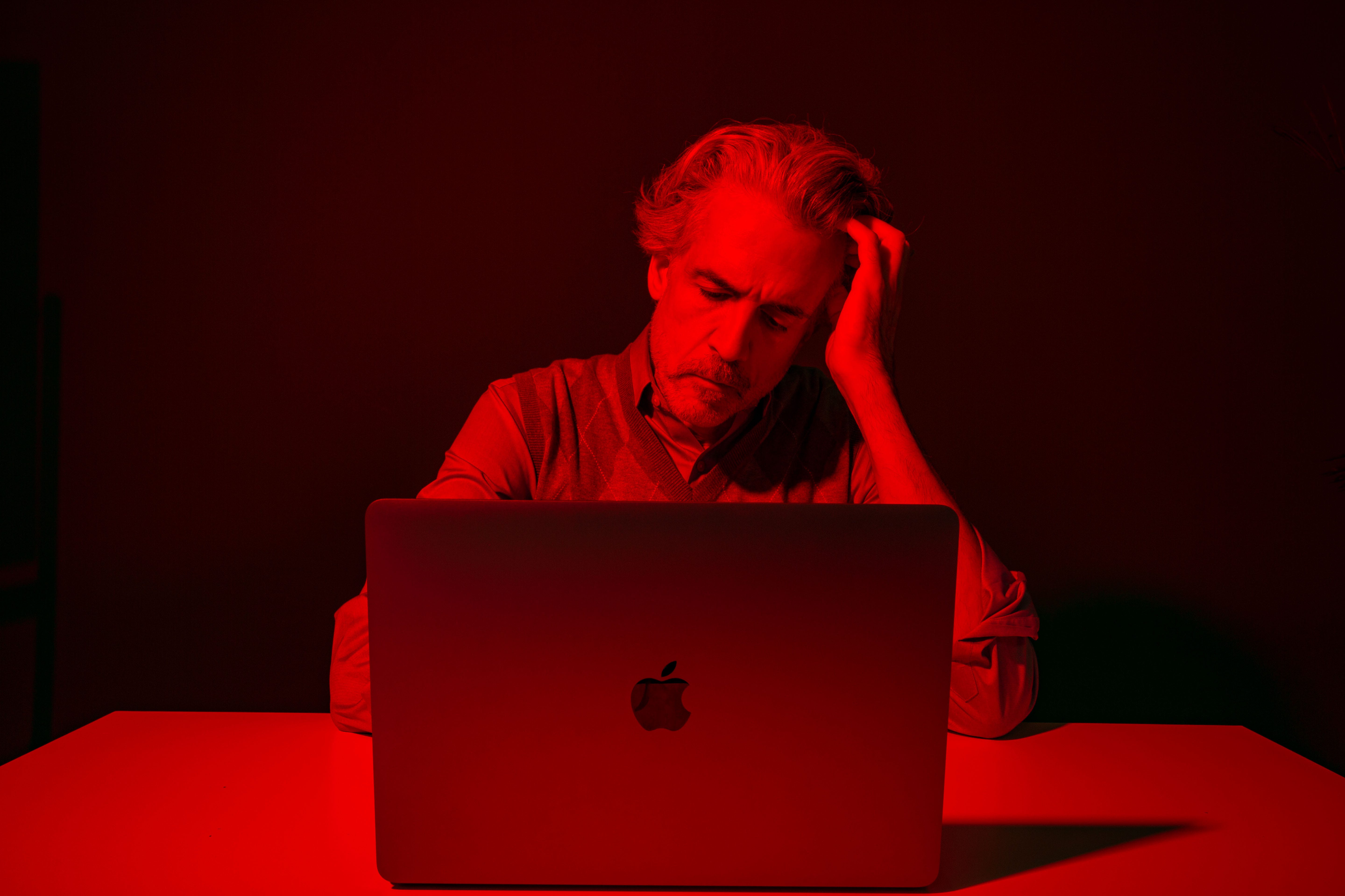 A contemplative and sad man sits at his laptop with a red hue.