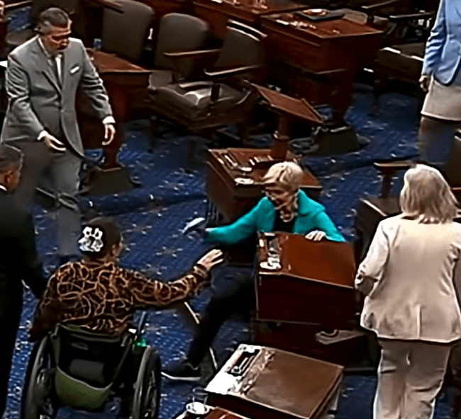 Screenshot of a chaotic scene in a legislative chamber with people interacting amidst overturned desks and chairs.