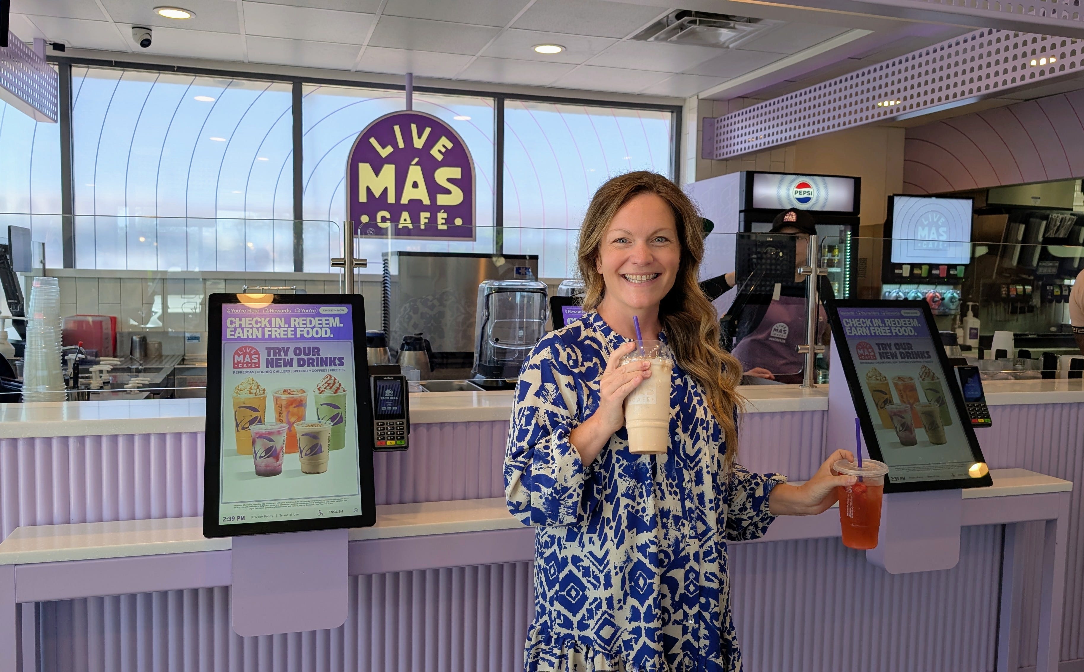 Author Michelle Stansbury smiling in Taco Bell Live Mas Cafe with drinks in each hand, ordering kiosks behind her