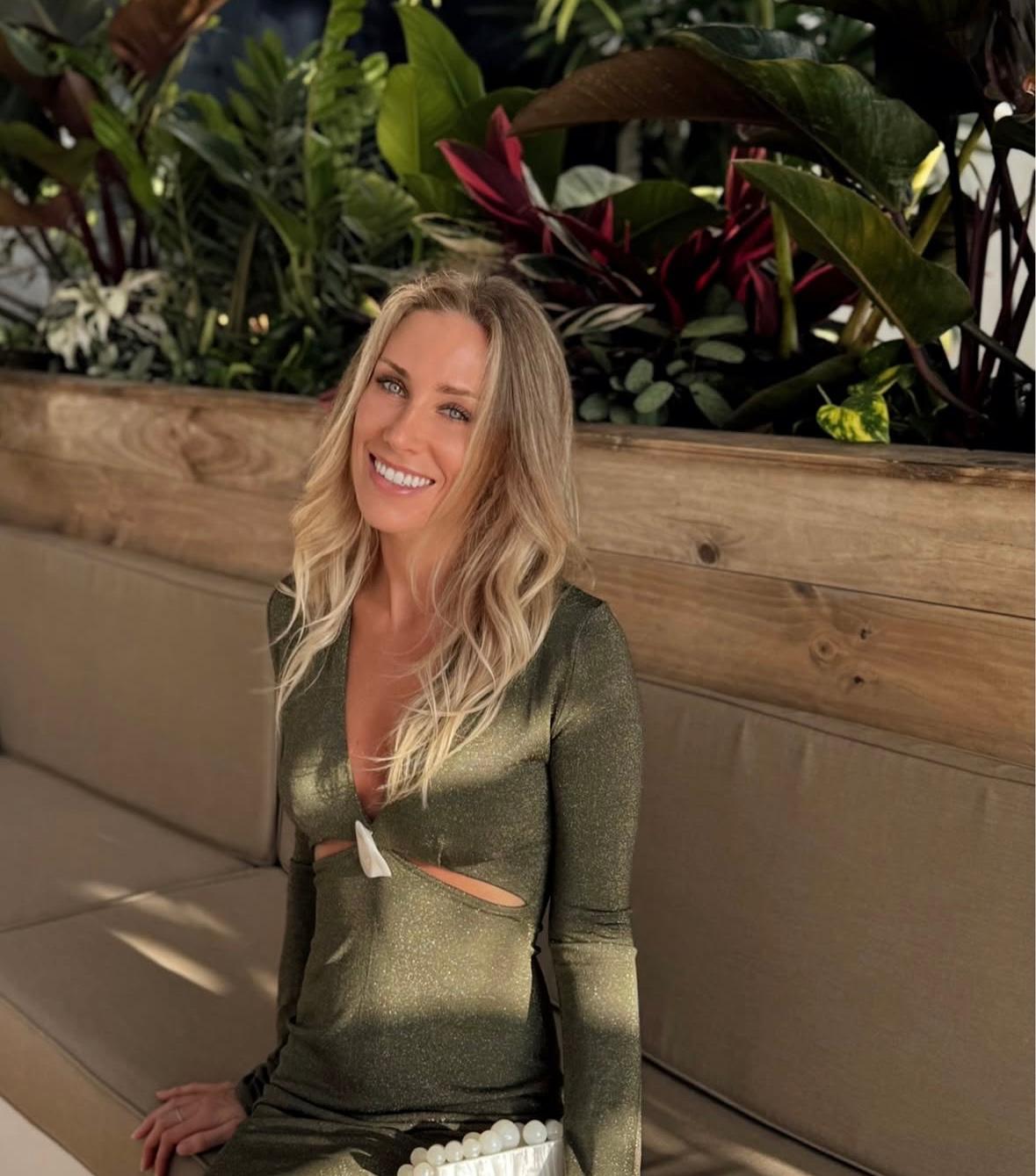 A woman smiles sitting on a padded bench with greenery in the background.