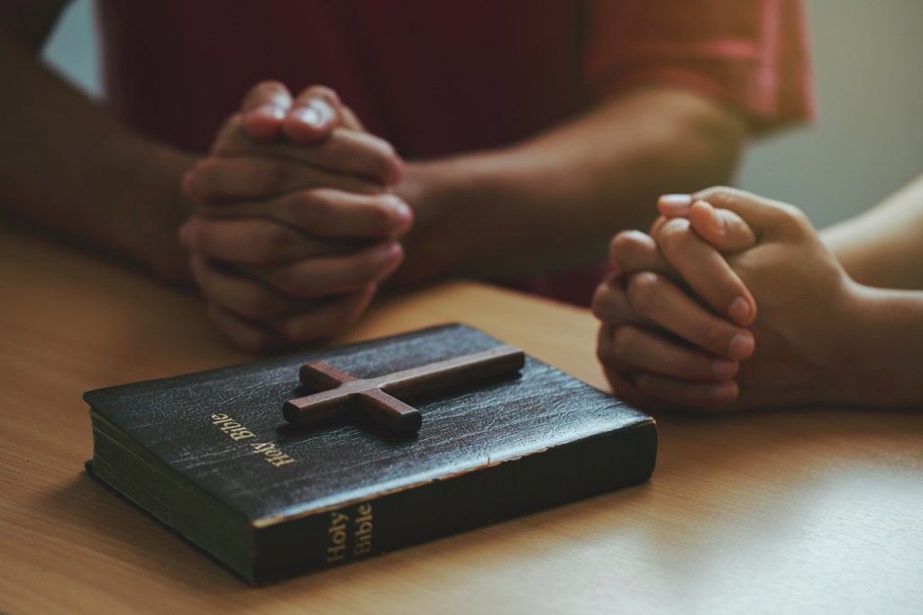 Two people praying with hands clasped over a Bible and wooden cross.