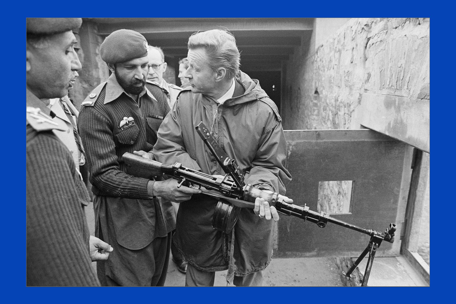 Brzezinski holds a machine gun as he talks to a soldier wearing a beret, also holding the gun. Another soldier is in the foreground with men behind them.