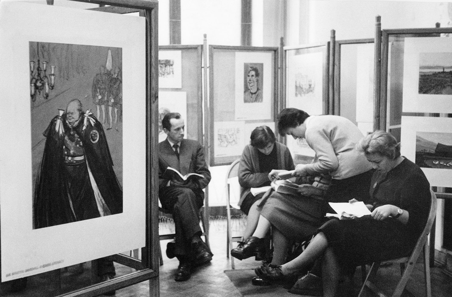 Four people sit on chairs holding books in a gallery-like space with paintings behind them.