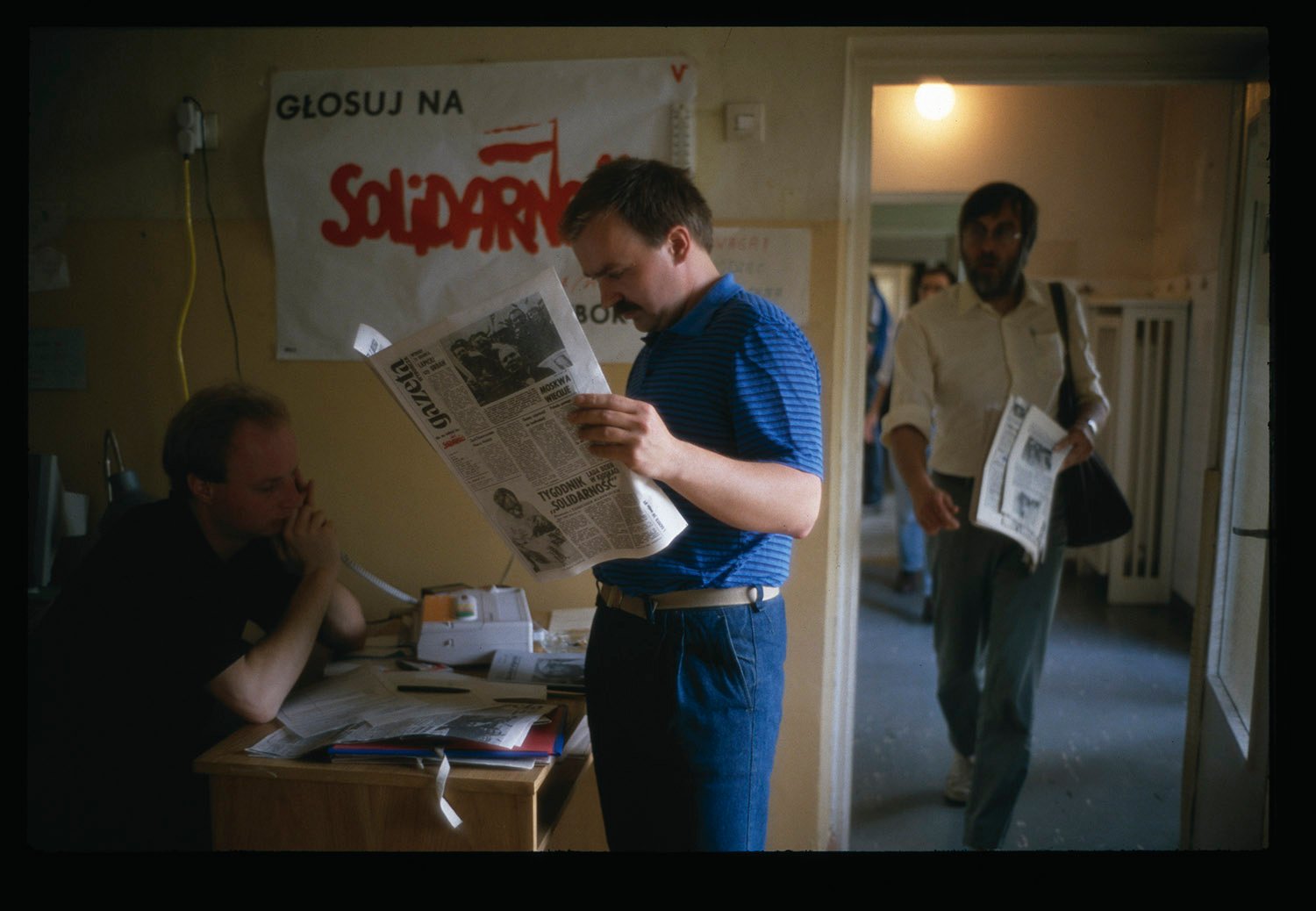 Two men hold newspapers in an office. The word Solidarity is seen in red on a sign on the wall.