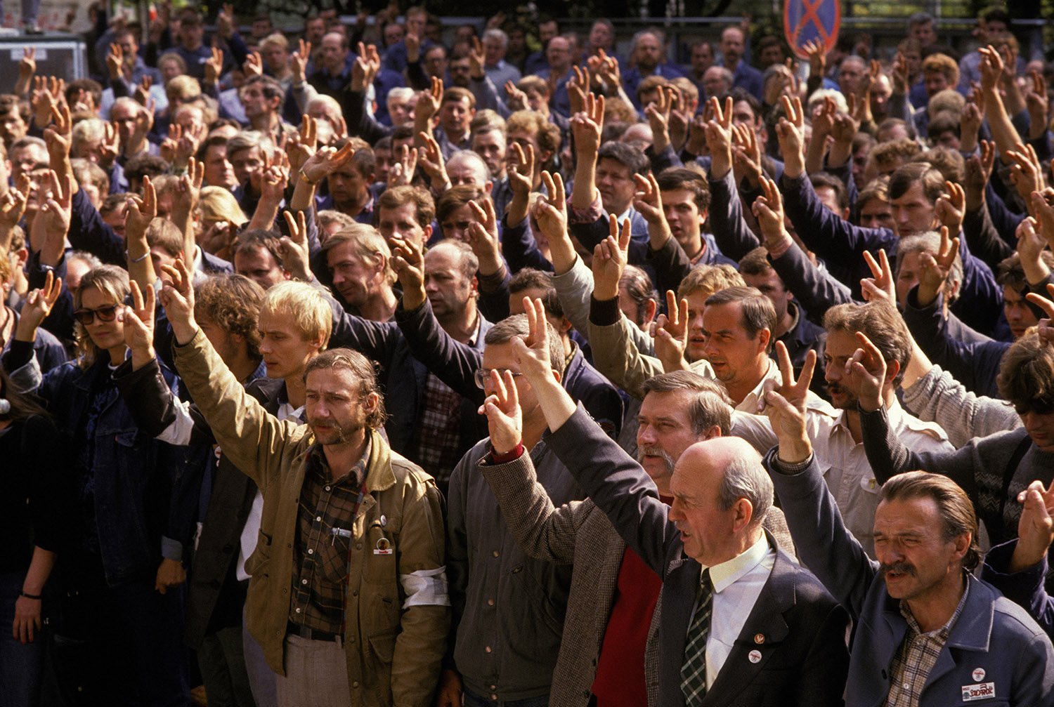 a large crowd of protesters stand with one arm outstretched with two fingers in a V sign.