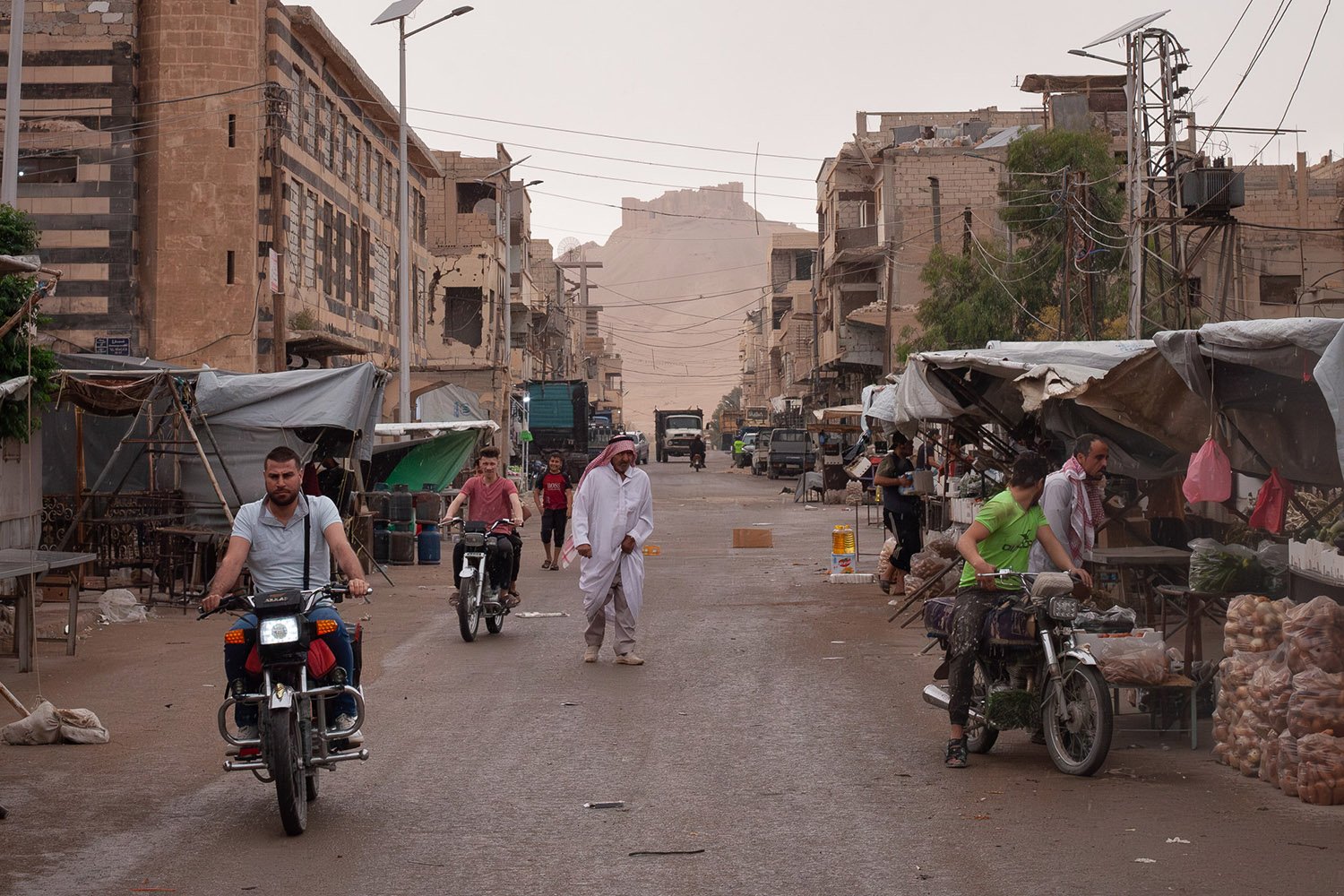 Men on motorbikes and others walking move along a street past stalls with awnings. Damaged buildings frame the street with a distant hill and fortress between.