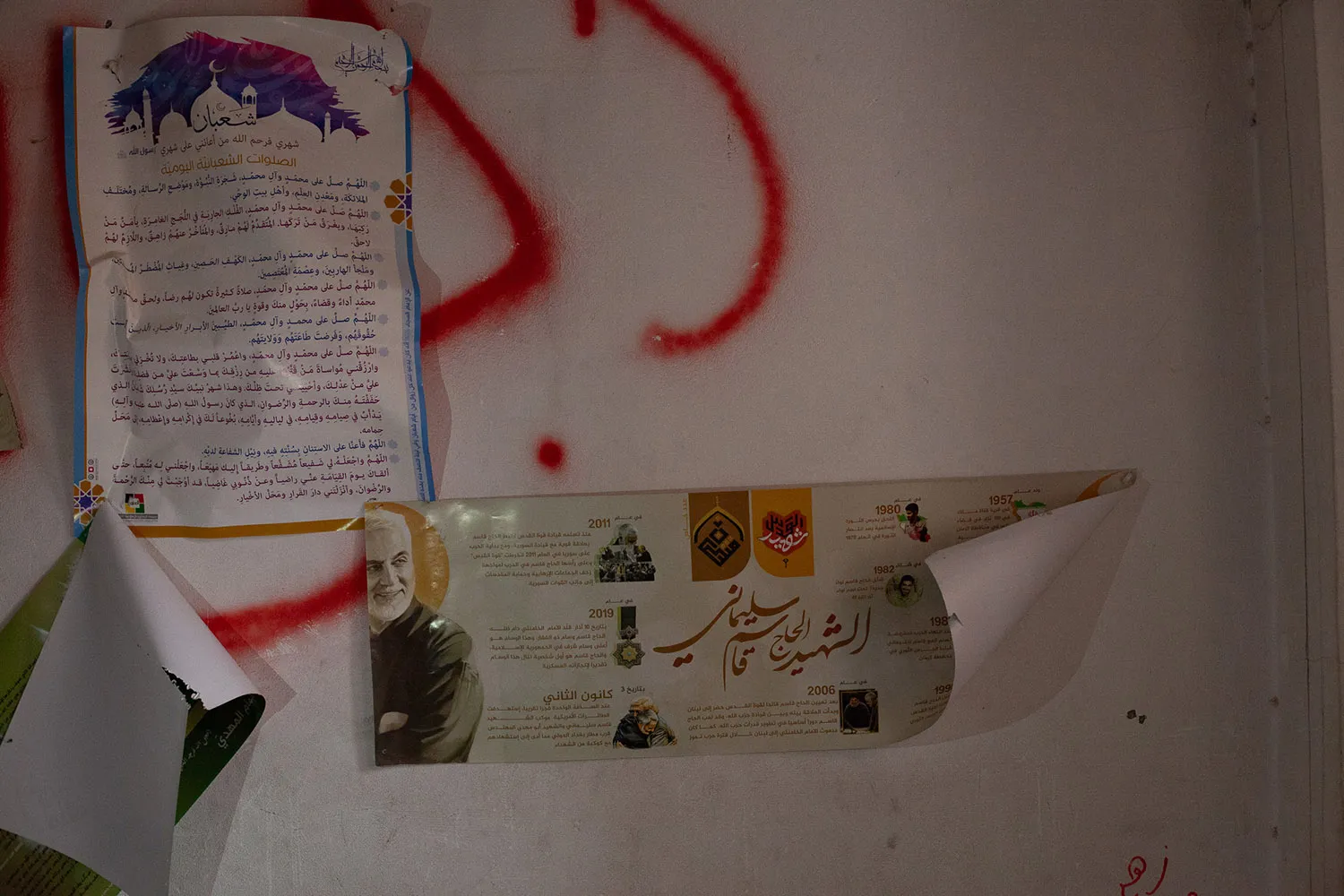 A wall with posters showing text in Arabic, red spray paint, and a picture of Suleimani.