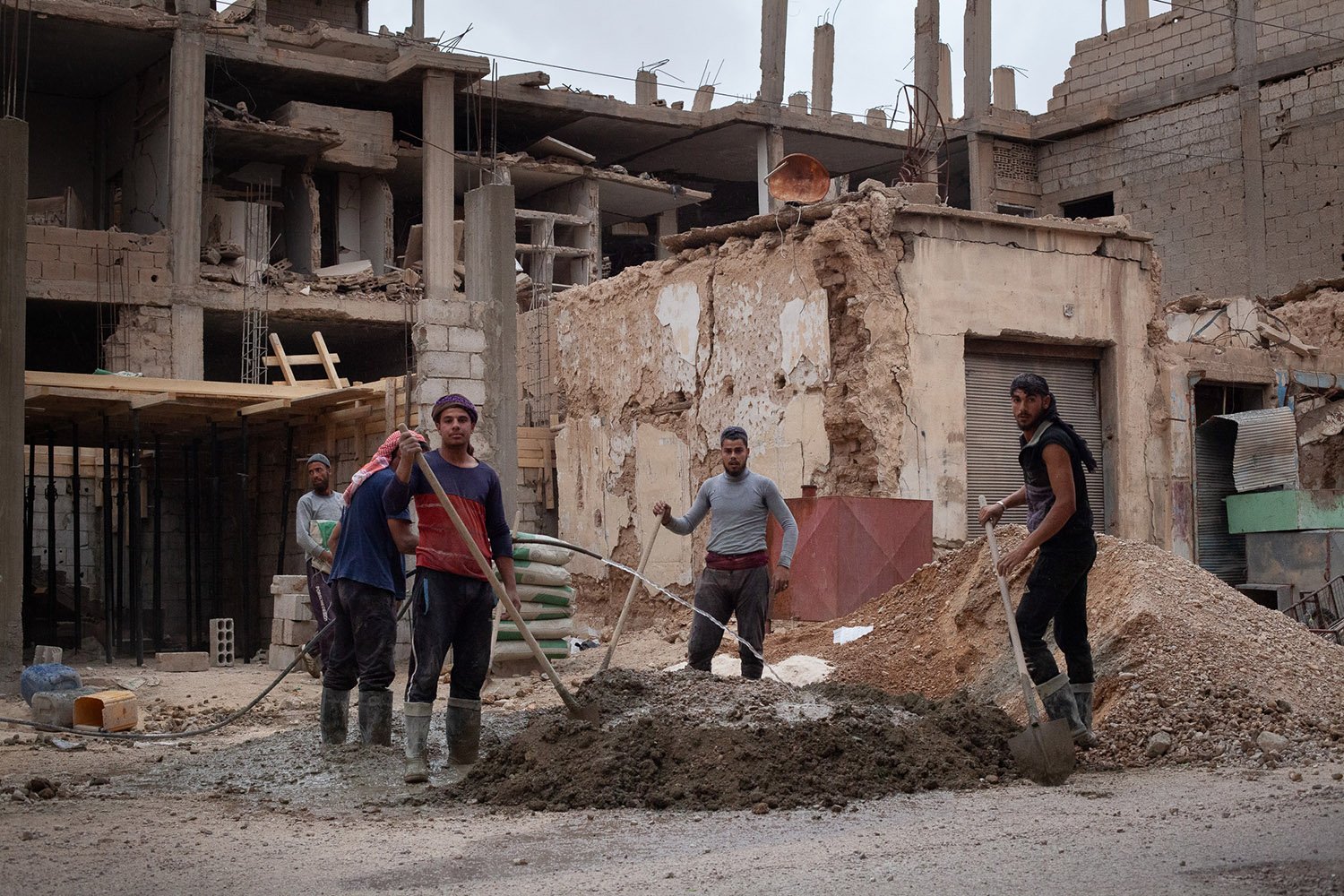 Five men hold shovels and other tools as they stand next to a pile of dirt or cement in front of heavily damaged buildings.