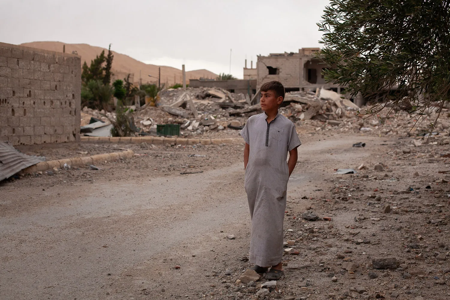 A boy with his hands in his pockets in a long robe on a dirt road covered with debris and damaged buildings behind.