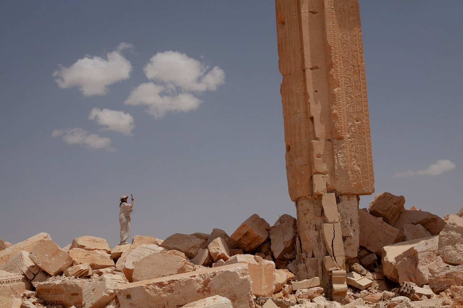A large column looms over a small person standing atop a large field of rubble holding up a phone to take a photo.