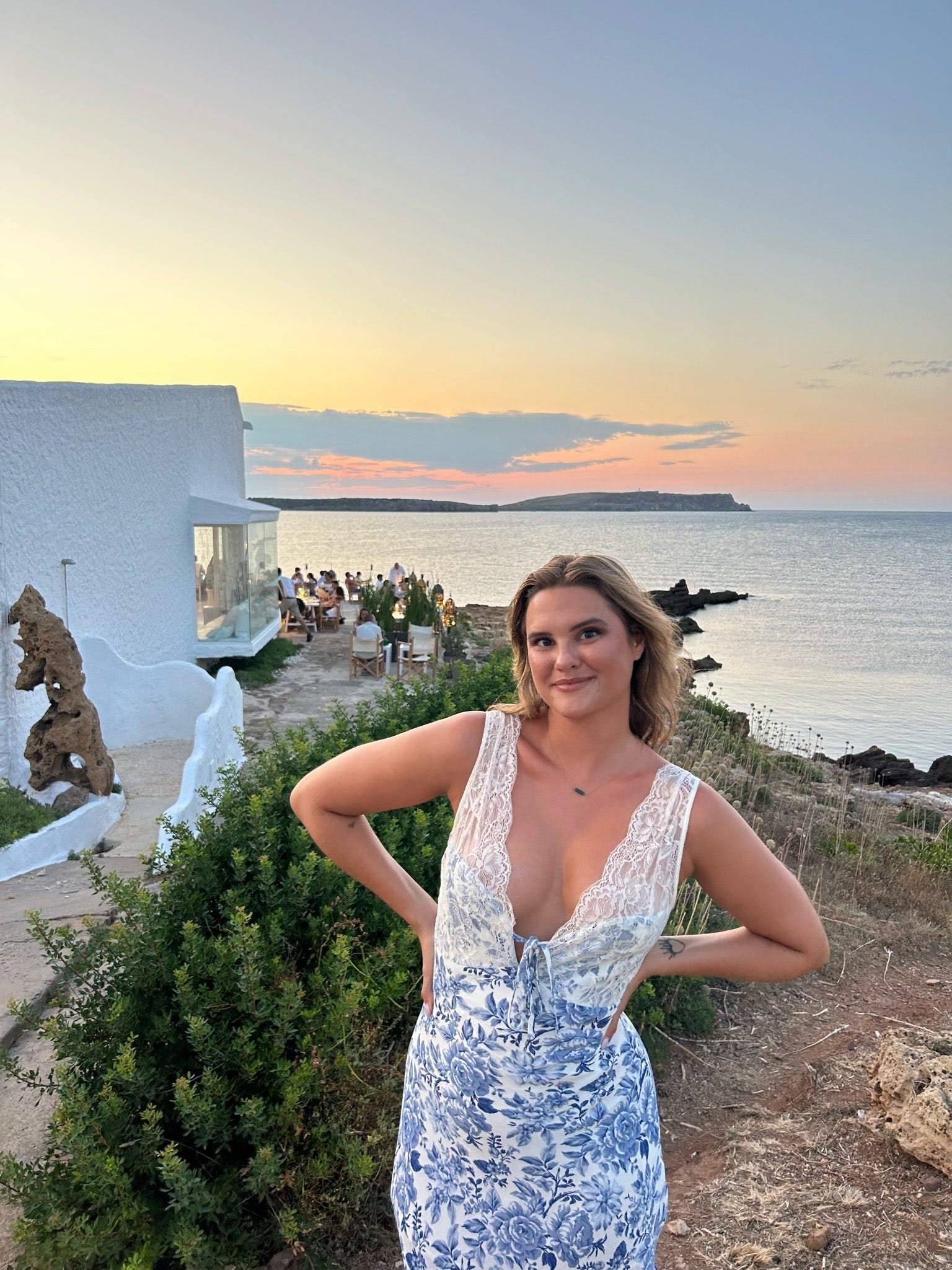 Parrie Hartley standing on a hill side overlooking the ocean, people sit at tables at a nearby resturant.