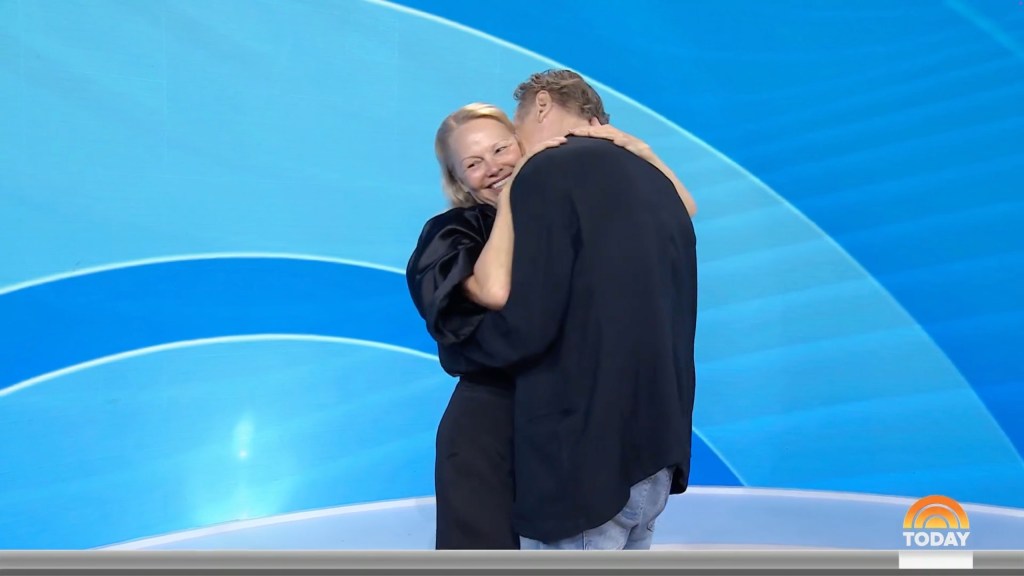 Pamela Anderson in black shirt and black pants, hugs Liam Neeson, wearing a black suit.