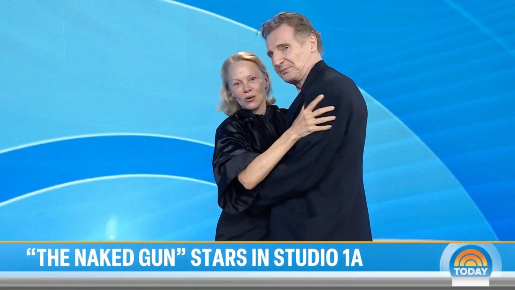Pamela Anderson in black shirt and black pants, hugs Liam Neeson, wearing a black suit.