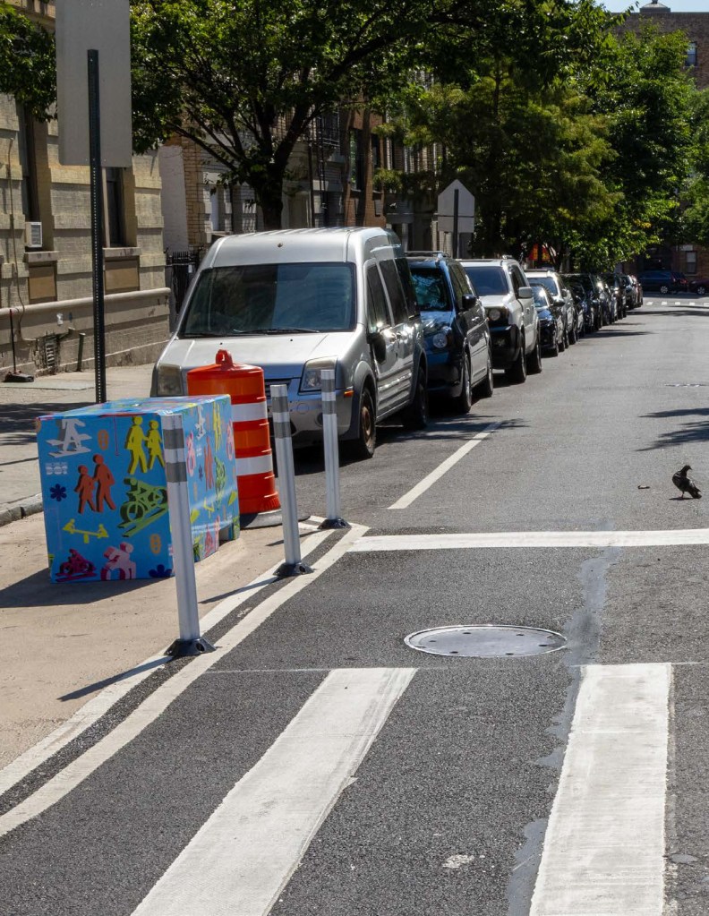 Daylighting installation at a city intersection with parked cars and decorated safety barriers.