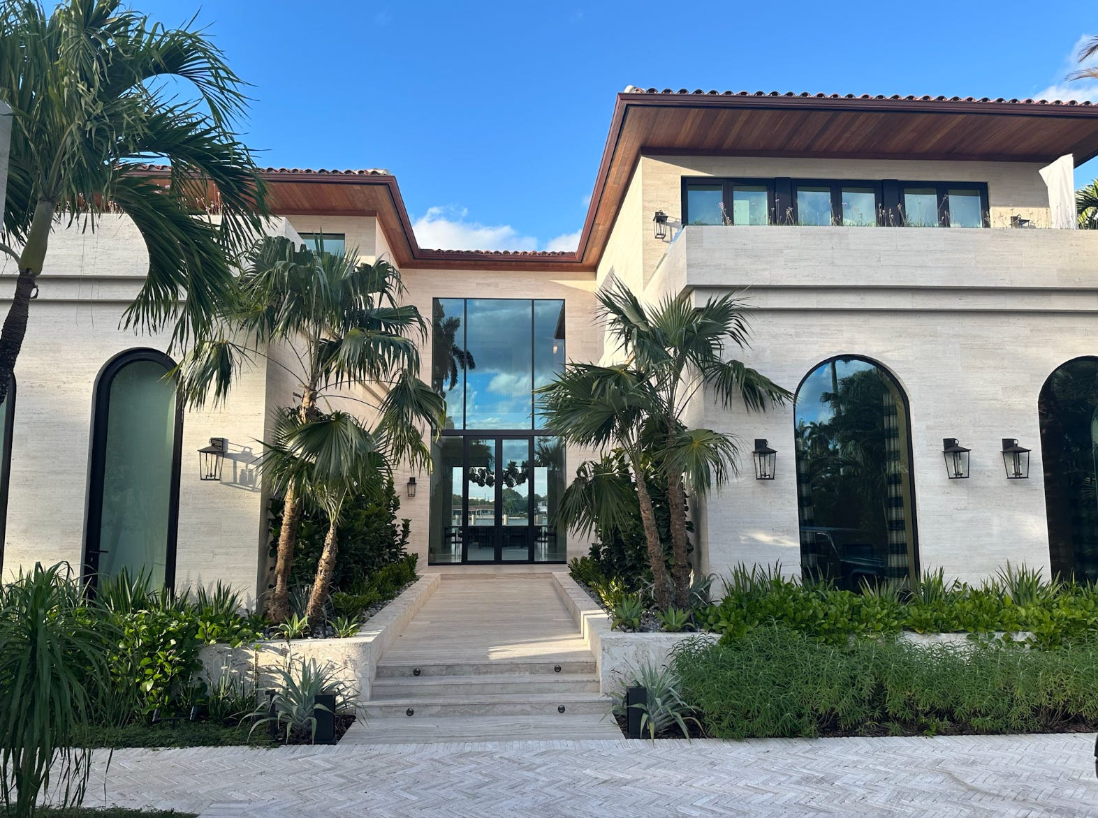 A front view of a Miami mansion with large windows, and palm trees lining the entrance.
