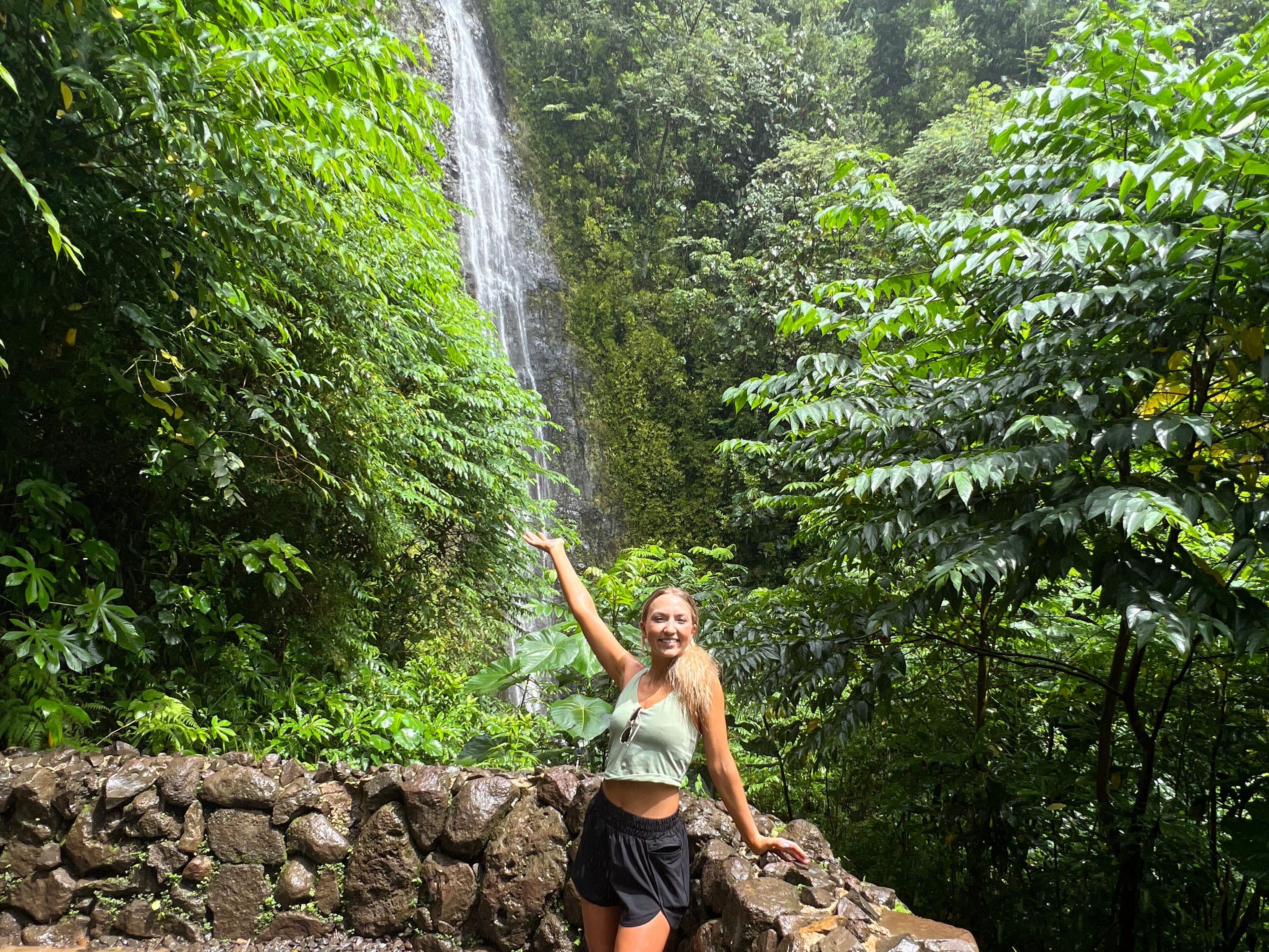 Chloe poses with her arm in the air in front of a waterfall surrounded by trees.