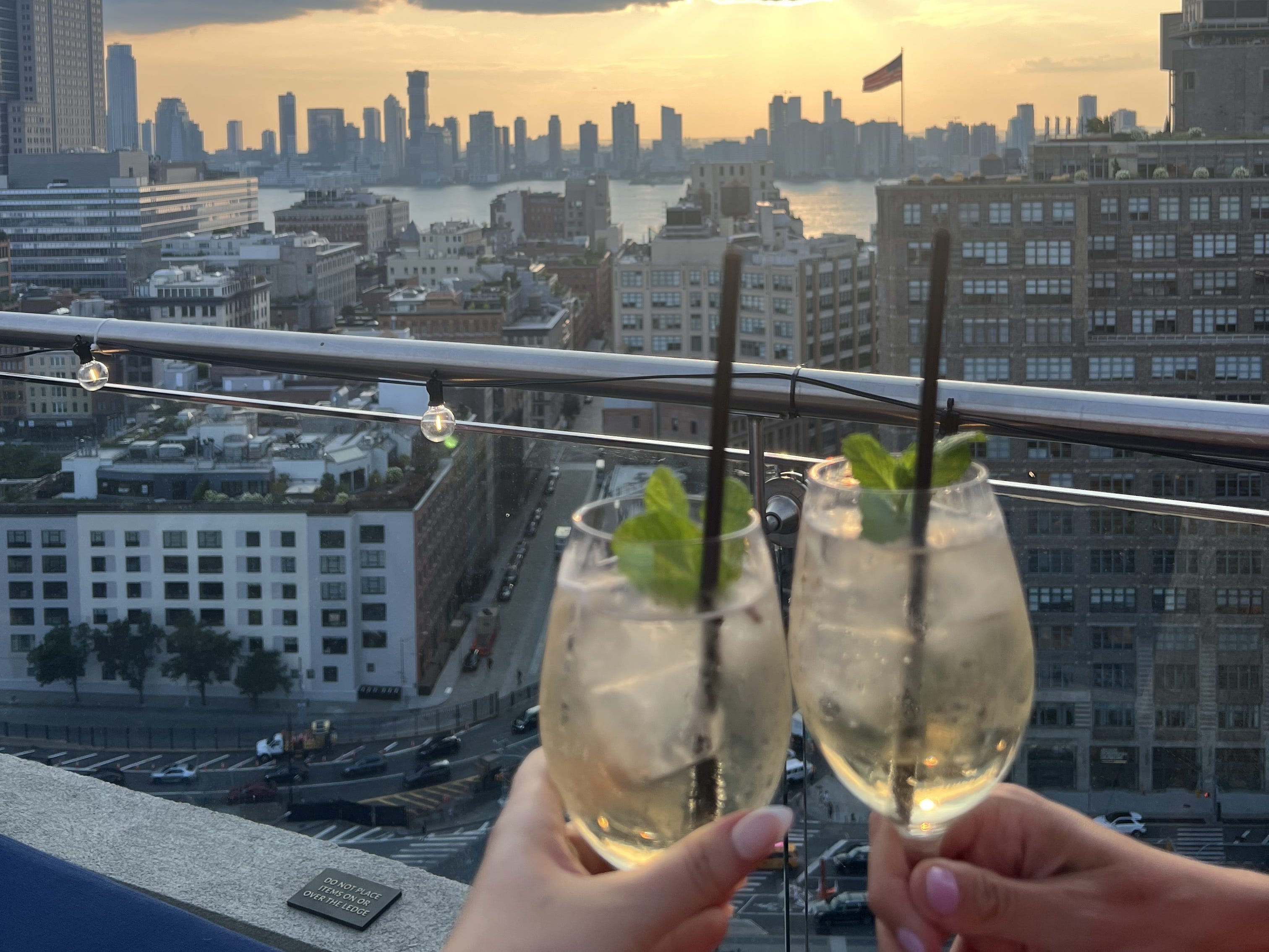 Two people holding cocktails on a rooftop in New York City during sunset.