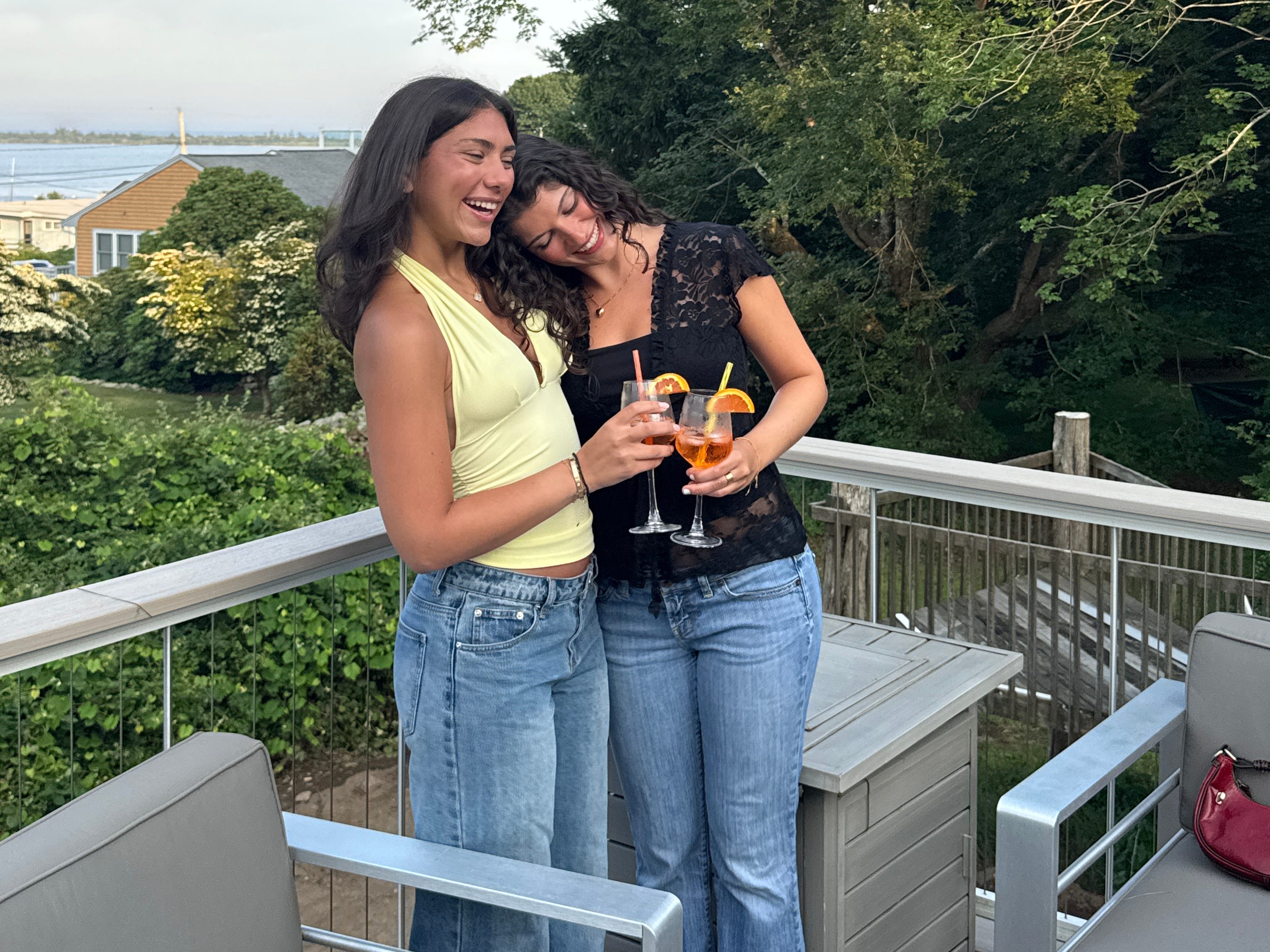 Emily and her friend hug while holding Aperol spritzes on a rooftop bar.