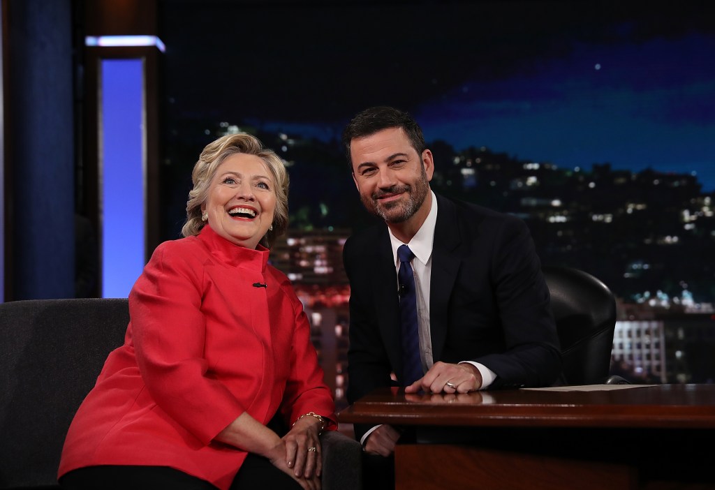 Hillary Clinton in a red jacket, Jimmy Kimmel in a black suit and black tie and white shirt.