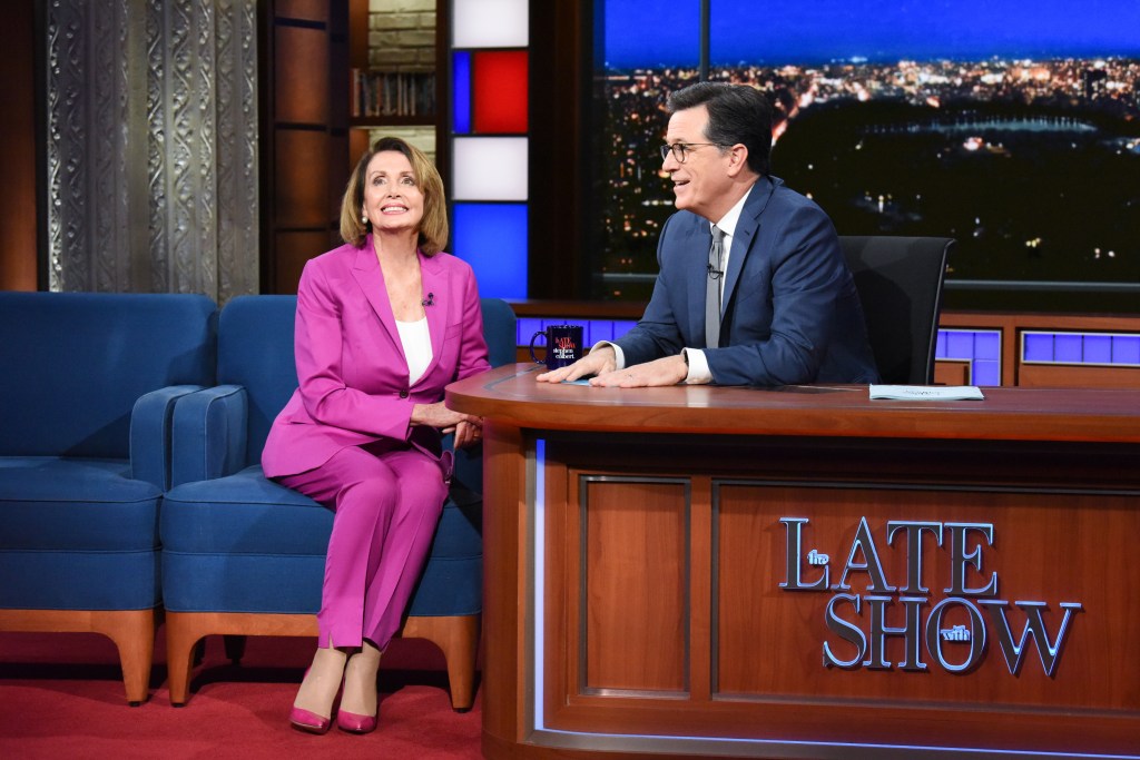 Rep. Nancy Pelosi in a hot pink pantsuit and pink heels, Steven Colbert in a blue suit and grey tie and white shirt.