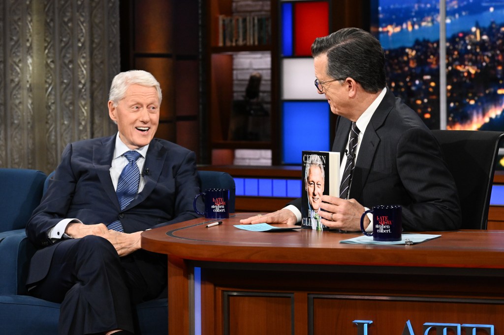 Bill Clinton in a navy suit, blue tie and blue shirt, Stephen Colbert in a dark grey suit, striped tie.