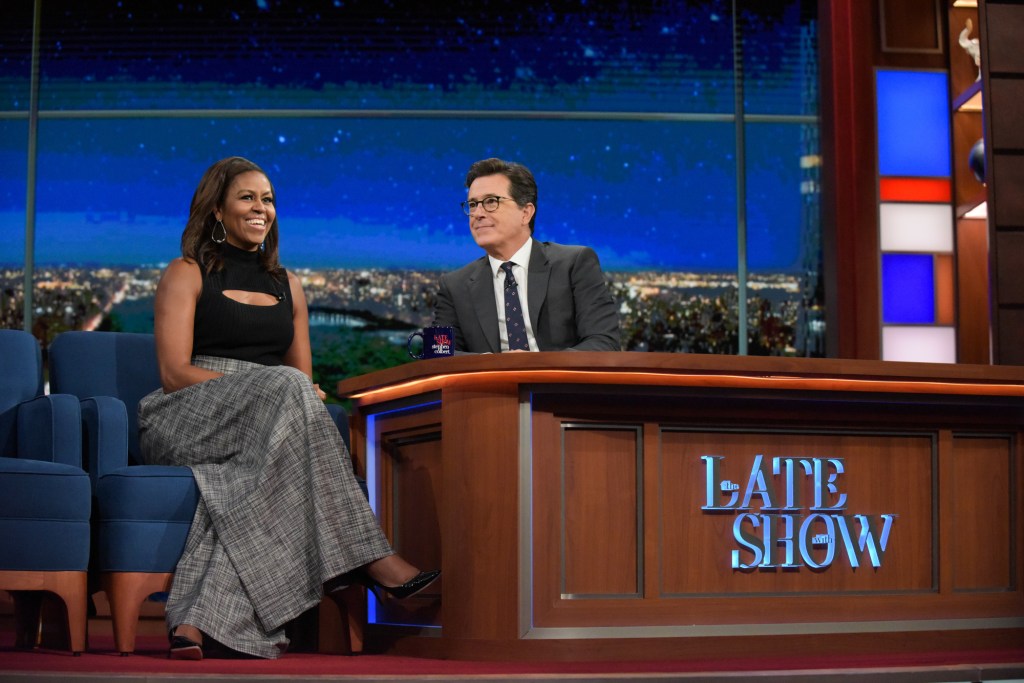 Michelle Obama in a black top and grey pants, Stephen Colbert in a grey suit.