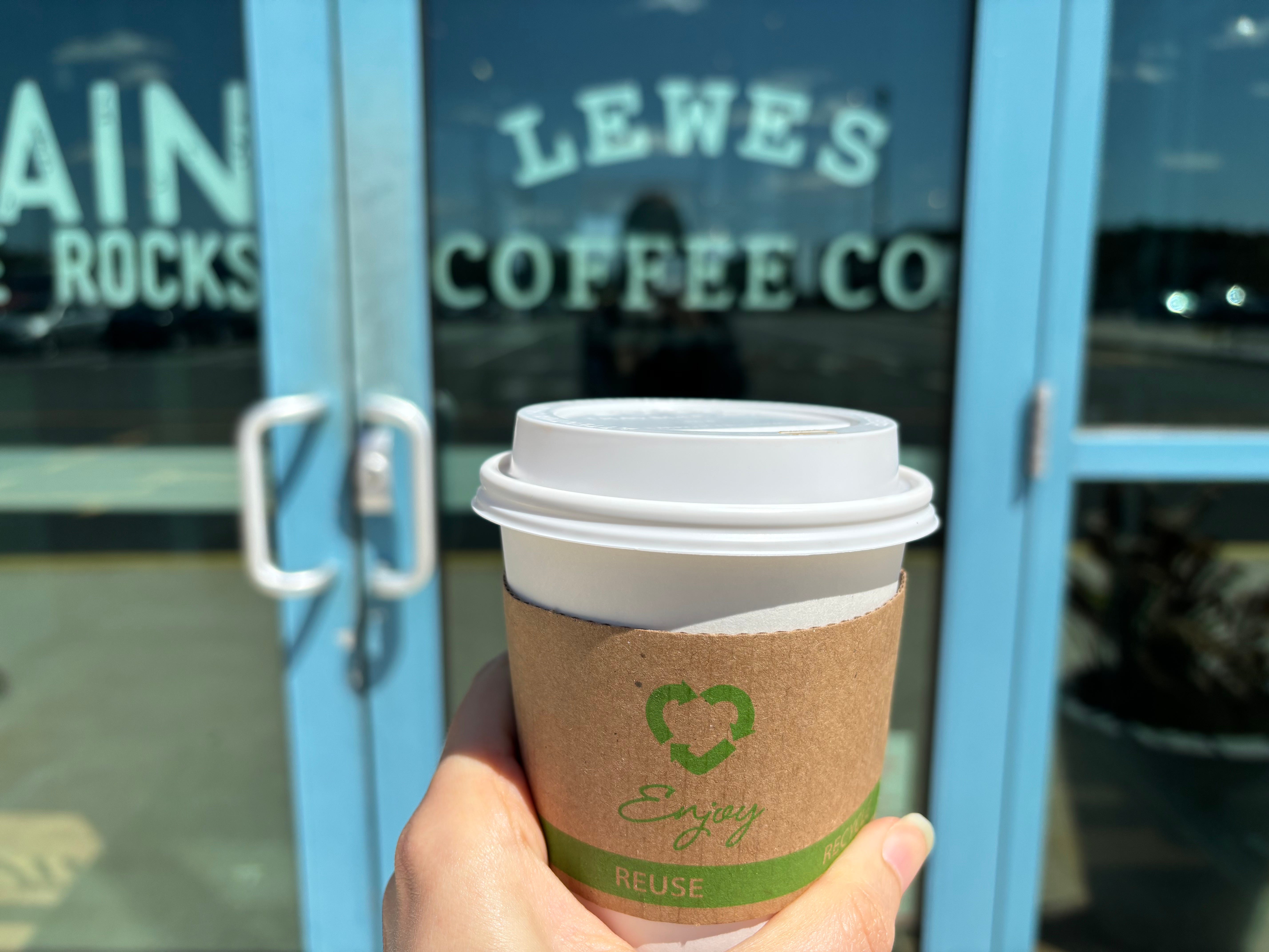 A hand holds a cup of coffee in front of a door that reads "Lewes Coffee Co."