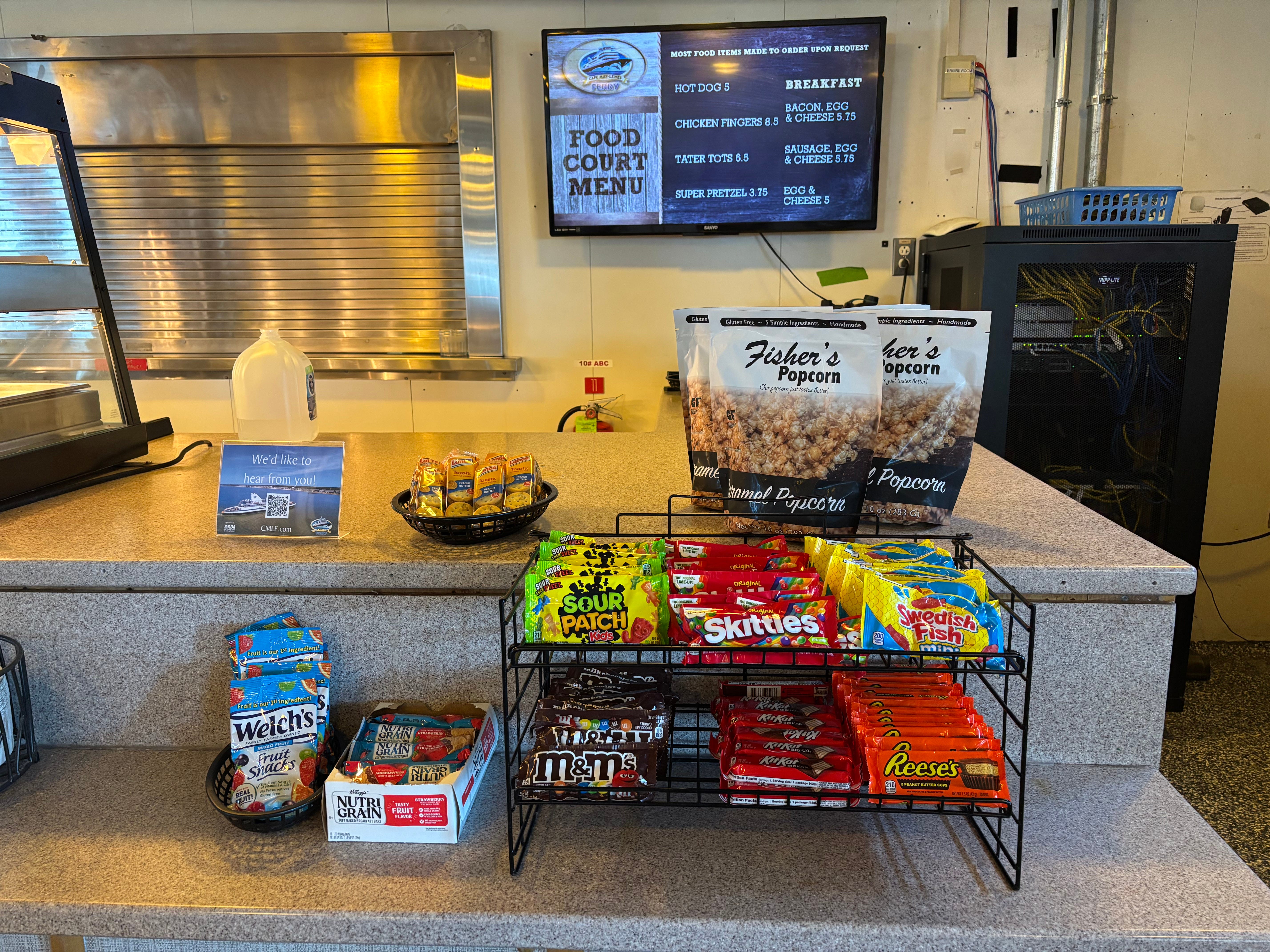 A snack stand on a ferry with popcorn, crackers, candy, and fruit snacks on the counter, and a digital sign on the wall listing other food options.