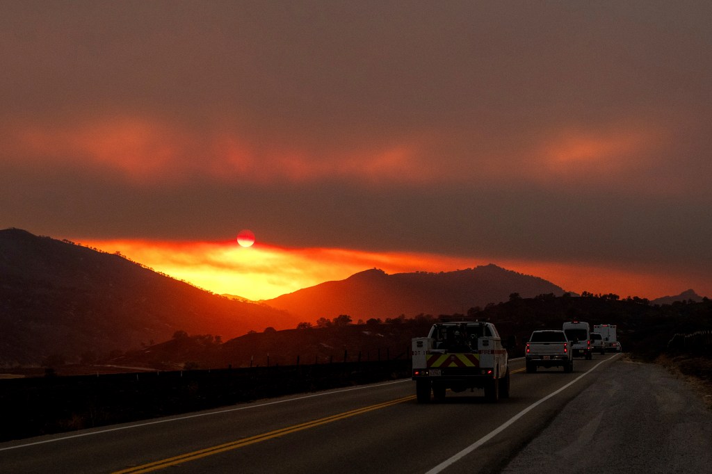 Sunset over fire-damaged hills in California, with fire trucks driving on a road.