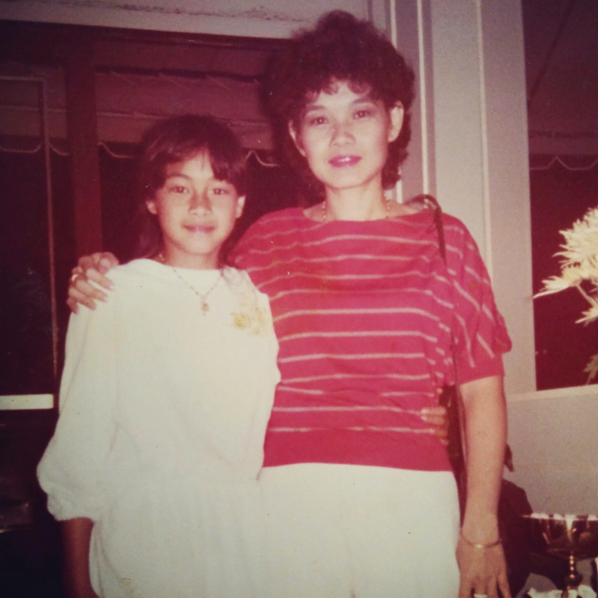 A young Metinee Kingpayome, who is dressed in white, posing in an old photo with her mother, who is dressed in a red and white striped shirt.