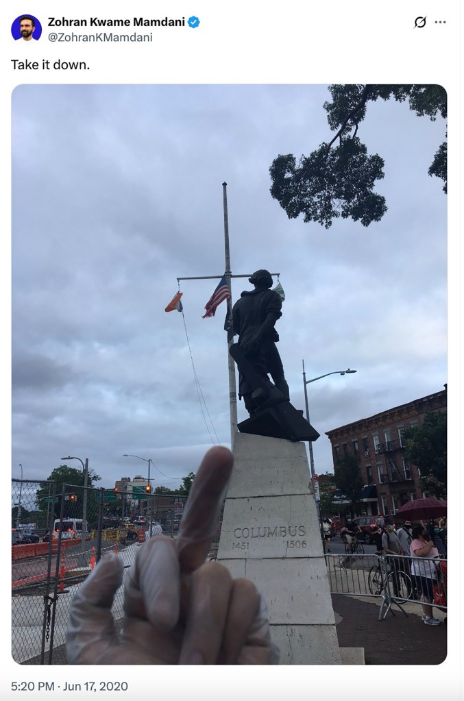 Photo of a Columbus statue with a middle finger in the foreground and a protest in the background.